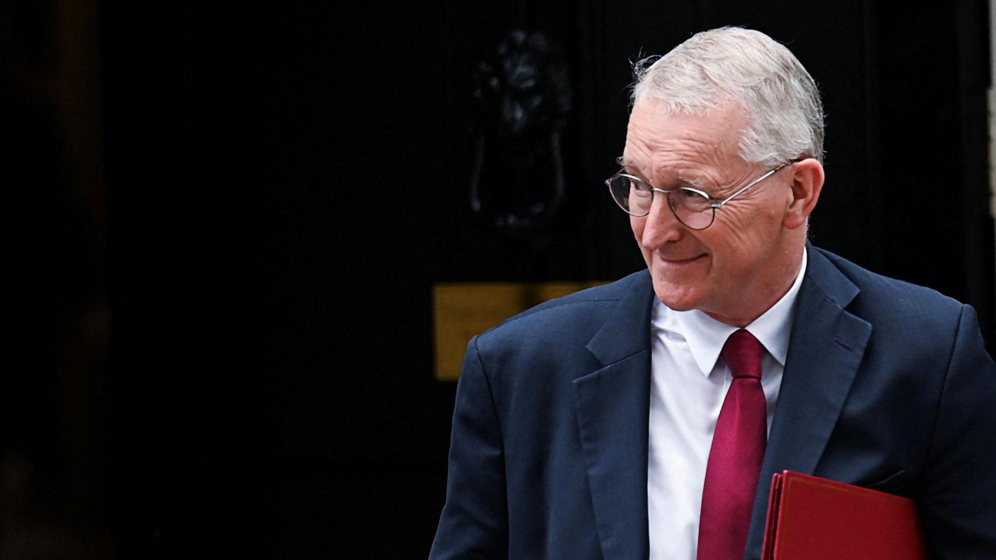 Secretary of State for Northern Ireland Hilary Benn, wearing a blue suit and a red tie. He has grey hair and round glasses. He is carrying a red file under one of his arms