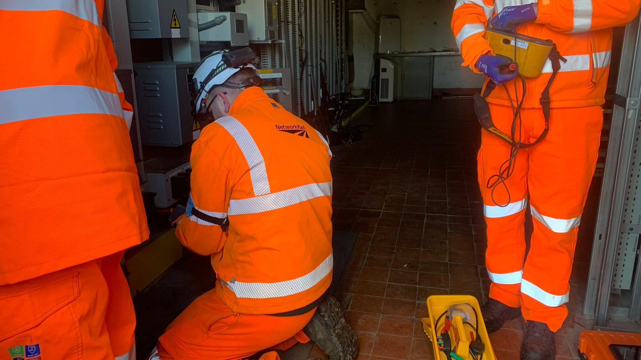 Three people wearing bright orange high-visibility uniforms are making repairs to wiring inside a room. One person is crouched down with their back to the camera while looking down at something in their hands. The other two people are standing.  