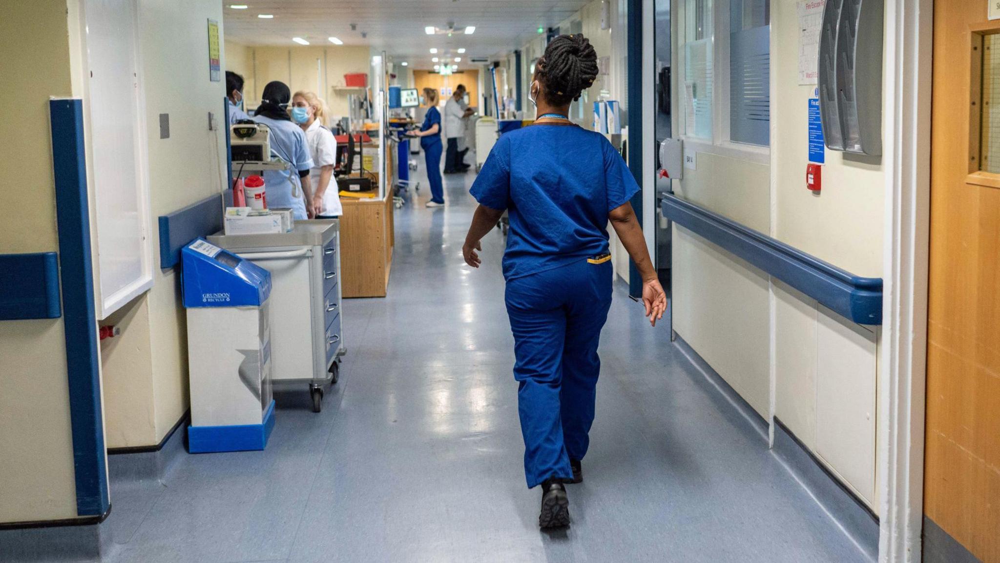 A nurse in blue scrubs walks down a hospital corridor.