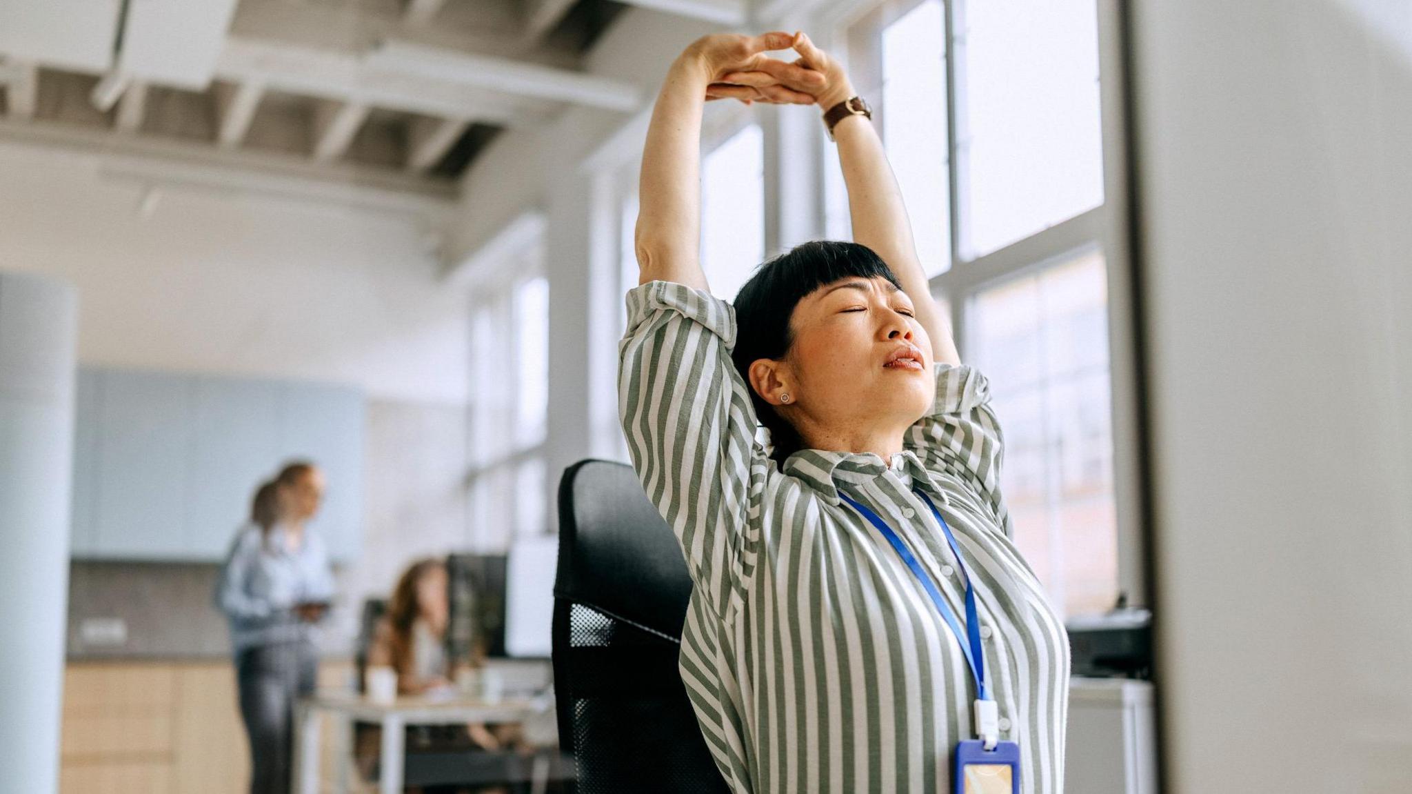 A busy woman takes a moment to stretch while working at her desk in a modern office, balancing productivity and well-being
