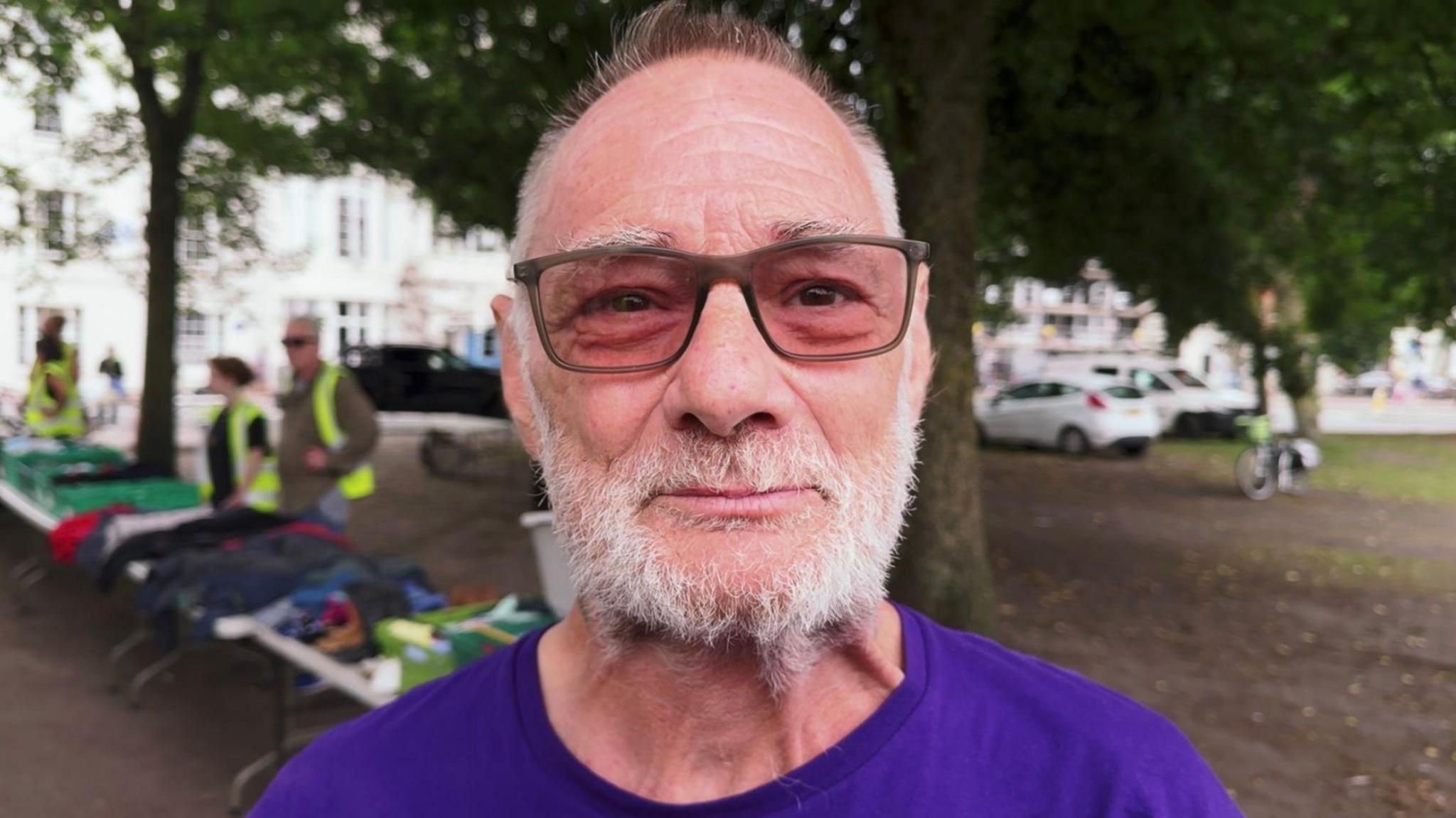 Alan, who has a white beard, wears glasses and a purple t-shirt. He is looking into the camera and standing in an outdoor area with trees behind him. The background is blurred but people in sleeveless hi-vis jackets can be seen standing behind trestle tables with clothing laid out on them