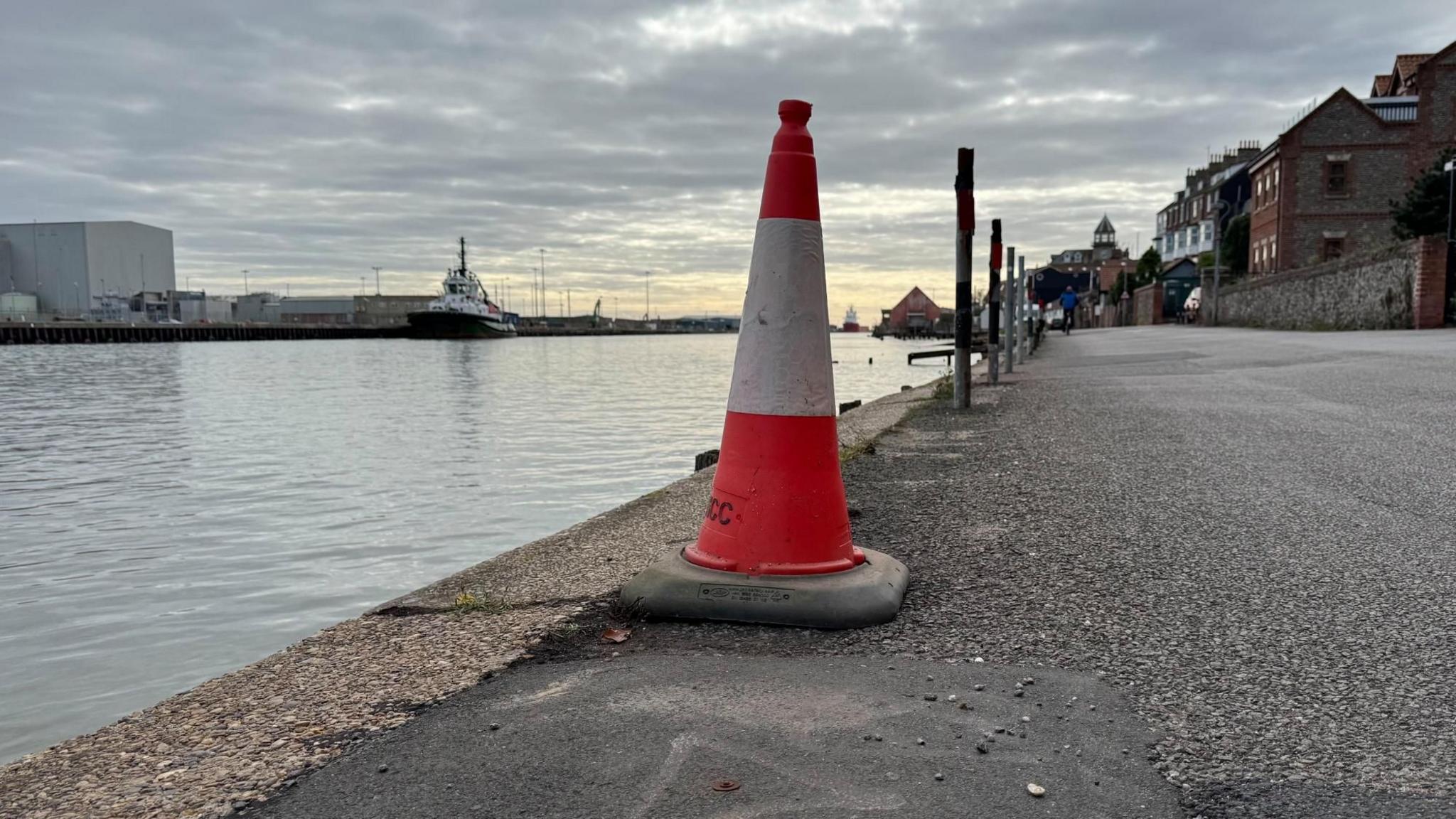 An orange and white traffic cone covering a small hole on the edge of a quayside road. The River Yare flows into the distance. There is housing on the Gorleston side of the river, and industrial buildings on the Great Yarmouth side.