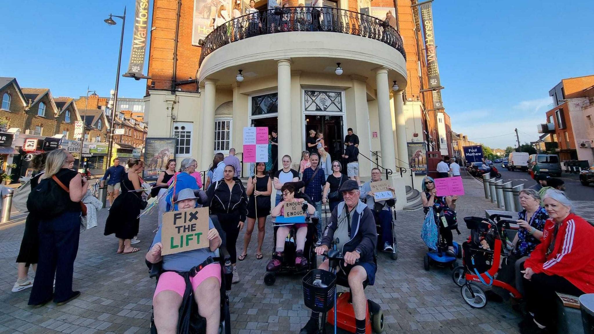 A group of protesters outside the theatre holding placards. At least six members of the protest group are in wheelchairs. 