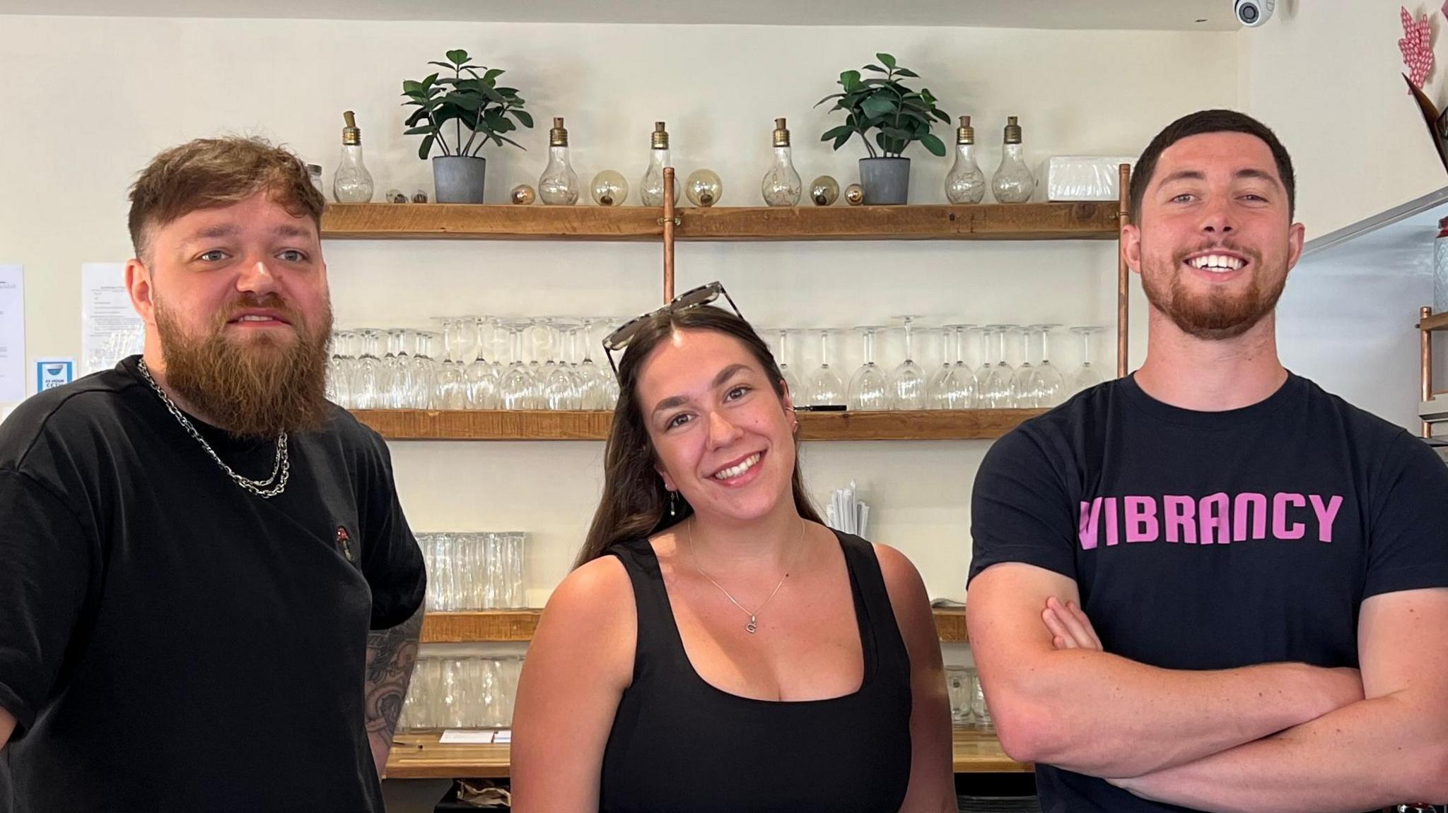 Mr Kabzinski, Ms Marsh and Mr Squire are next to each other in that order. They are stood behind a bar at the citizen hub. Mr Kabzinski has one arm on the counter and the other on his hip. He is wearing a black top and silver chain. He has brown hair and a long brown beard. Ms Marsh is smiling at the camera while wearing a black top, earrings and silver necklace. She has long brown hair and sunglasses on top of her head. Mr Squire is also smiling at the camera. He has his arms folded across his chest and is wearing a black top with 'Vibrancy' written on the front in pink. He has brown hair and a beard. Behind the three are shelves with wine glasses