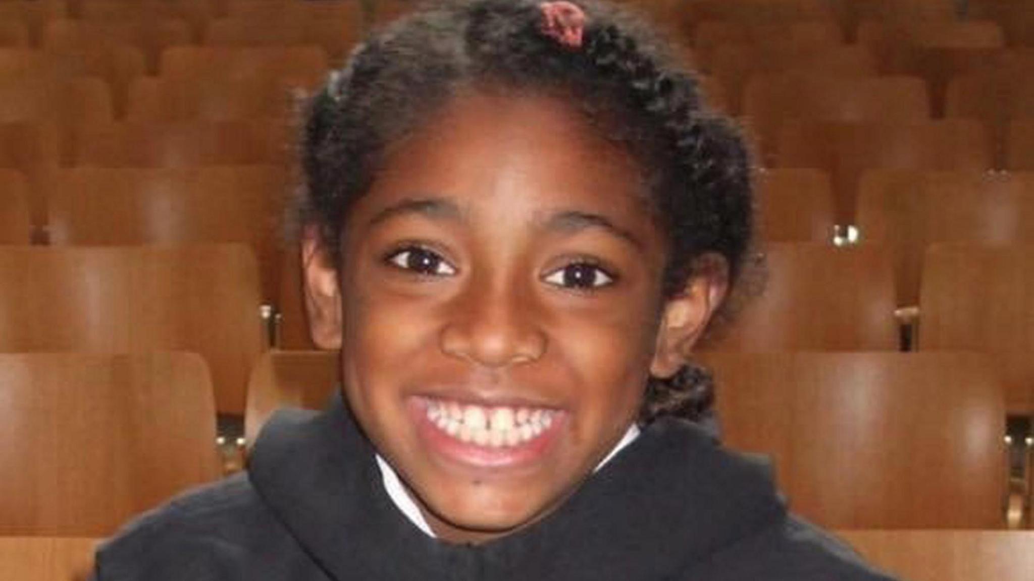  Ella Adoo-Kissi-Debrah with big smile, wearing a black jumper with rows of chairs in the background