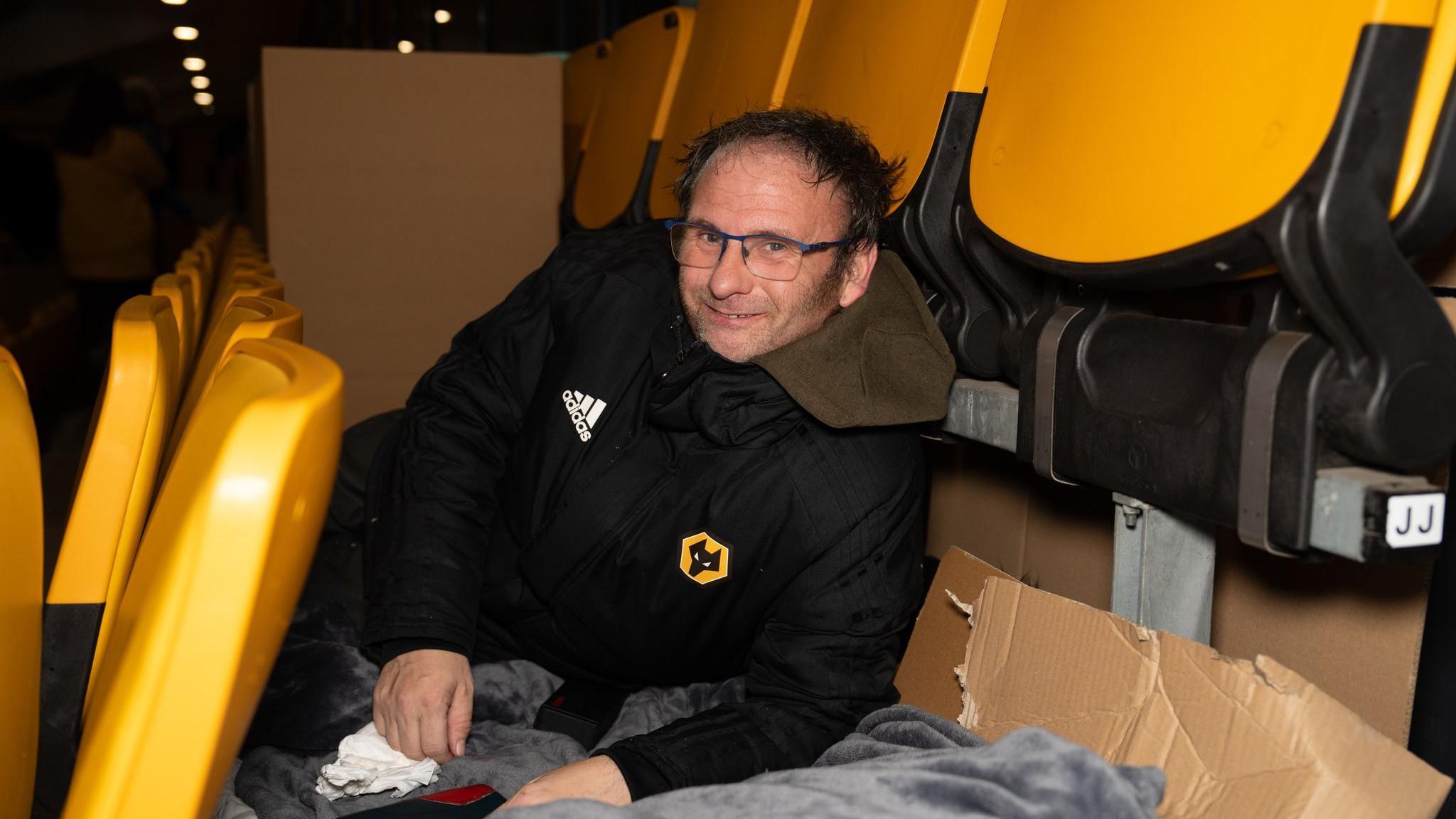 A man lies on the floor between seats in a stadium, on cardboard with blankets laid on top. The man is wearing a black jacket with the Wolves yellow and black logo on it and has dark brown hair and glasses. It is dark but the stand is lit.