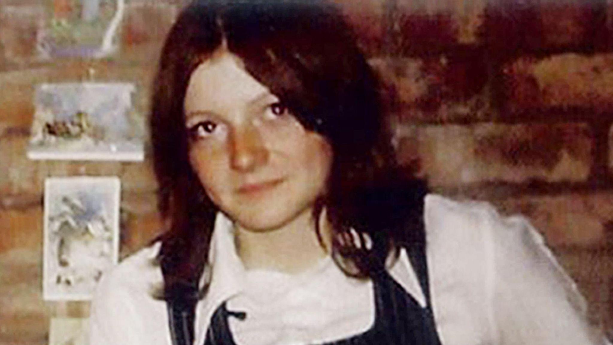 An old snapshot of a young woman with shoulder-length brown hair in a centre parting, a white shirt with pointed 70's style collar and a denim pinafore dress. She is inside against an exposed brick wall.