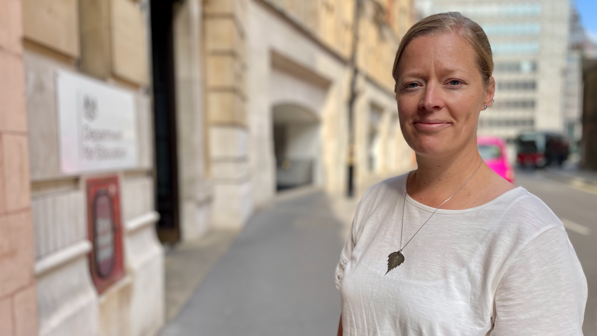 Rebecca Larkin wears a white T-shirt with a silver necklace. It is a sunny day and she stands on the street outside the Department for Education in London.