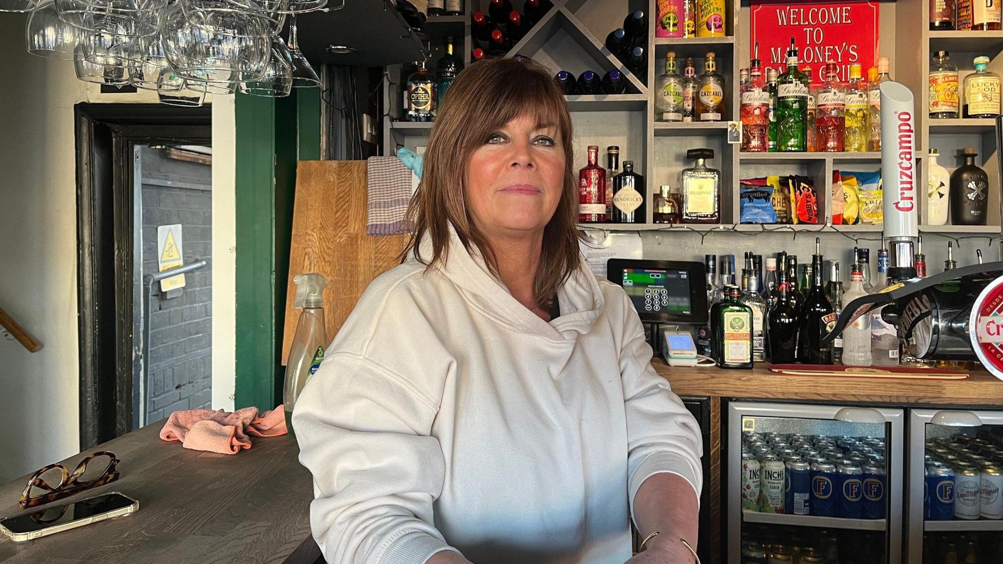 Louise Maloney sitting behind the bar in her pub. Wine glasses hang down from the bar above and there are bottles of spirits and beers behind her, as well as two fridges full of cans. Louise looks at the camera with a half smile, she wears a white hooded top and has shoulder length brown hair with a fringe.