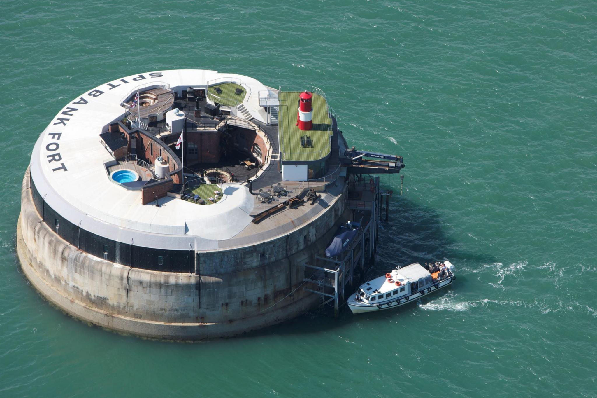 Spitbank Fort, a circular construction in the sea off Portsmouth. It is grey and its name is painted on the top. It has a little red lighthouse on one side of its surrounding wall and a swimming pool can be seen from the time it was a hotel. A Union Jack flies.