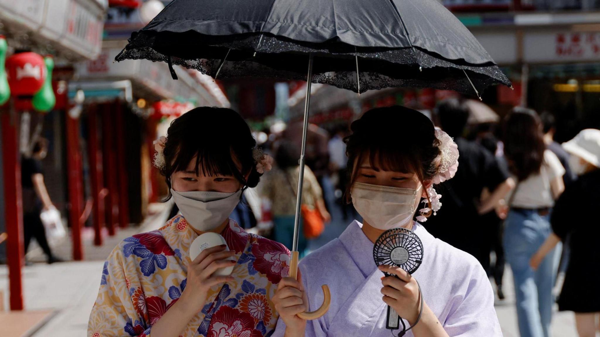 Two women, dressed in summer kimonos, hold an umbrella and portable fans as they walk through a Tokyo street.