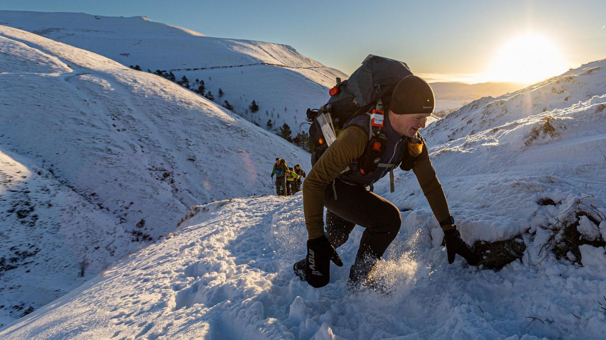 A man dressed in black with a rucksack climbing up a steep snow-covered path, with hills and other runners in the distance