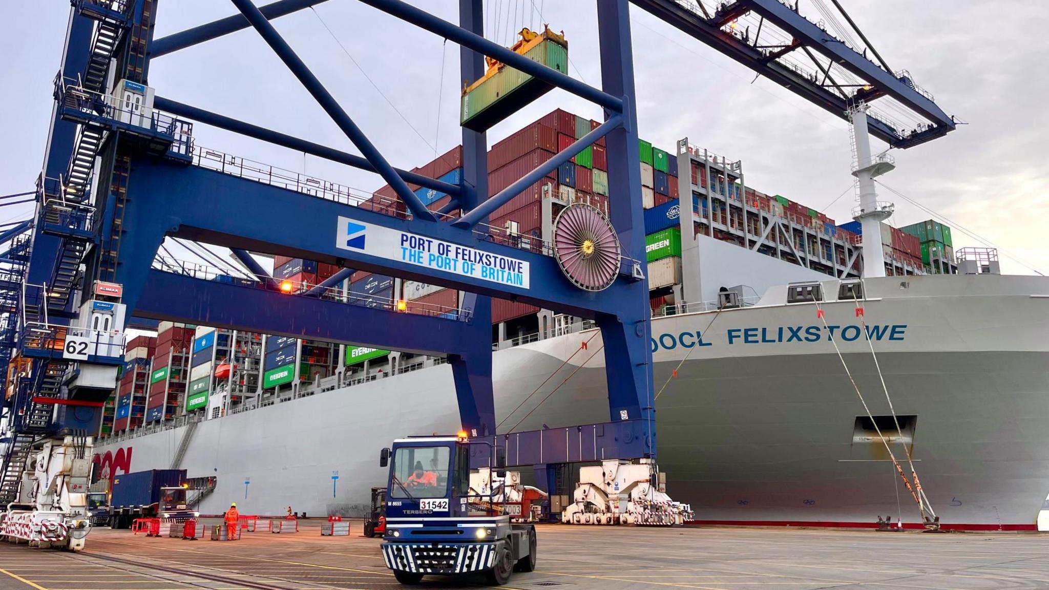 A small port vehicle is dwarfed by a crane that bears Port of Felixstowe branding. In the background there is a huge container ship, its deck packed with different coloured containers.