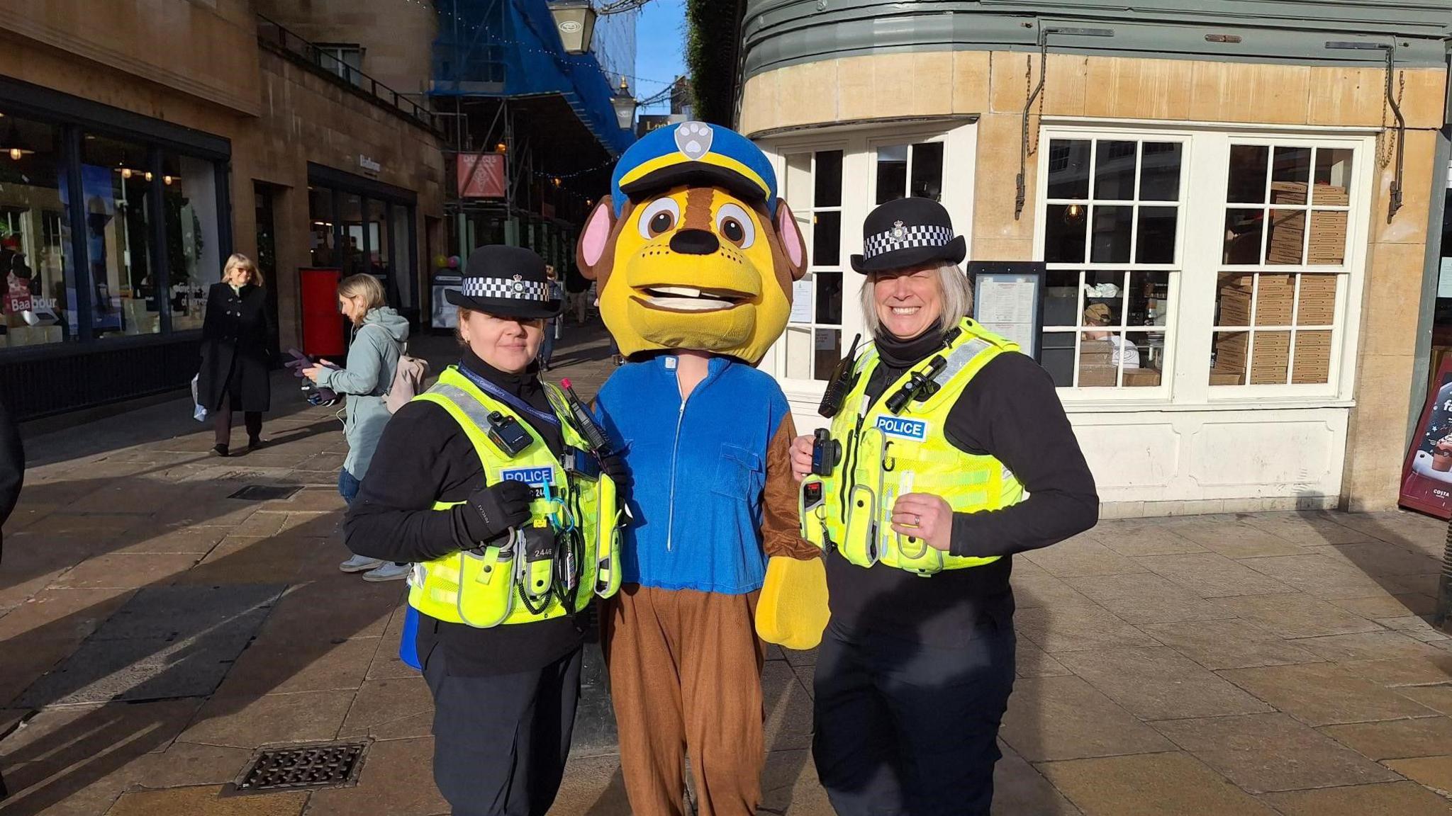 Two police women wearing their high-visibility green jacket and black hats and black full-sleeve shirts and trousers - standing on either side of a brown monkey mascot in Cambridge city centre 