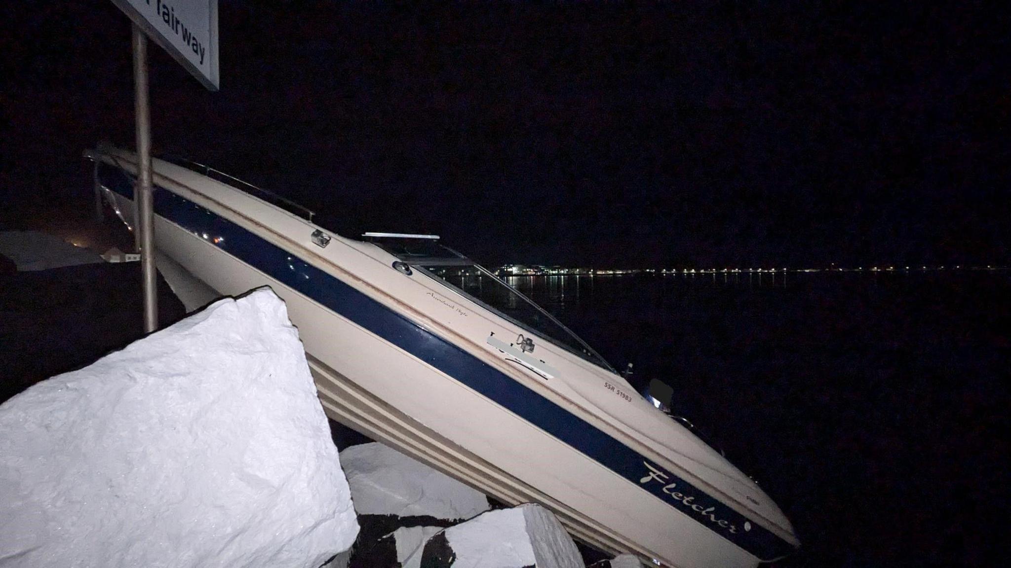 A small white and blue motorboat aground at the entrance to the Queen Elizabeth II Marina. The small vessel is at an angle on some white rocks.