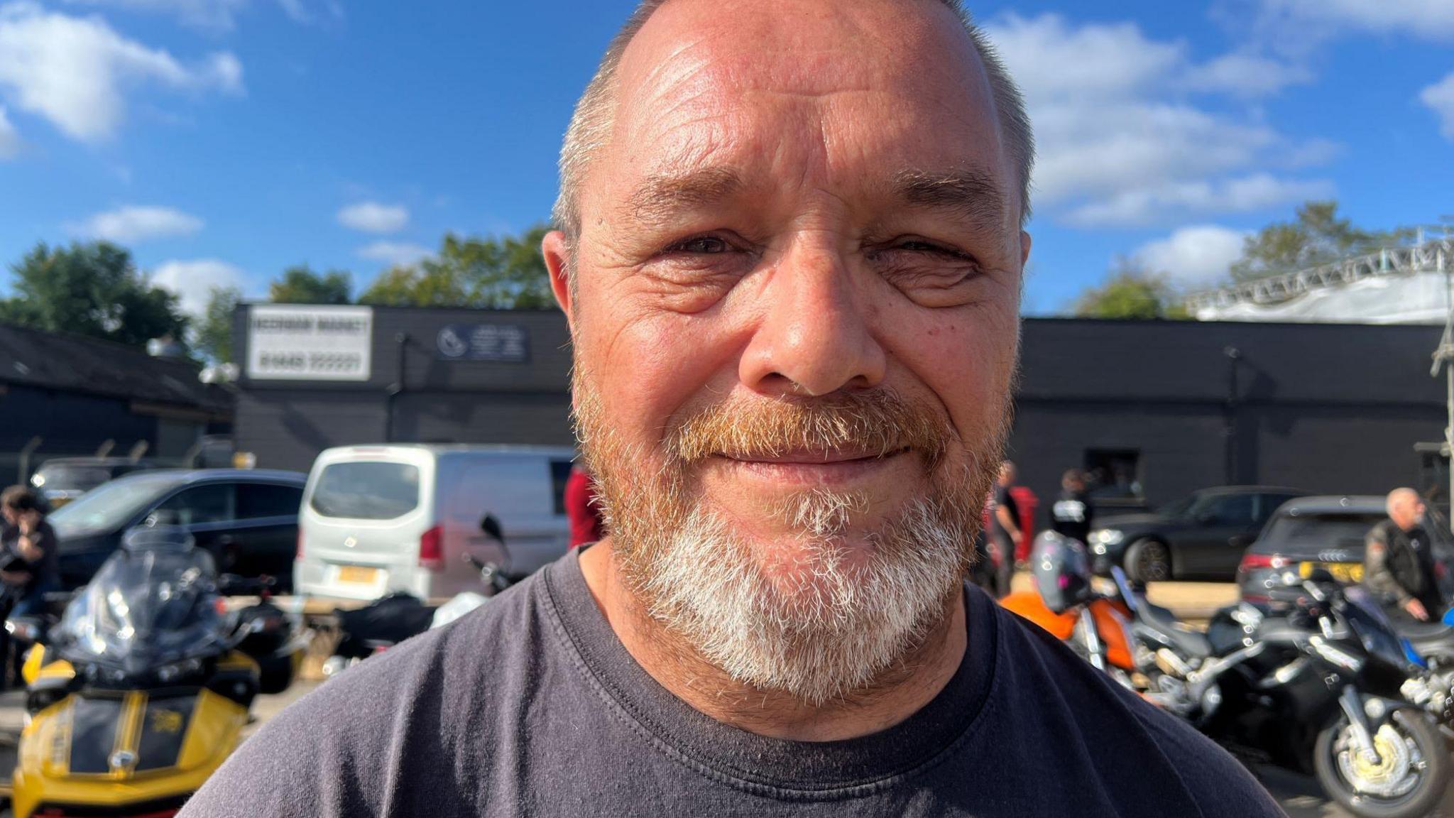 A man with a beard looking at the camera and smiling. There are motorbikes parked behind him.