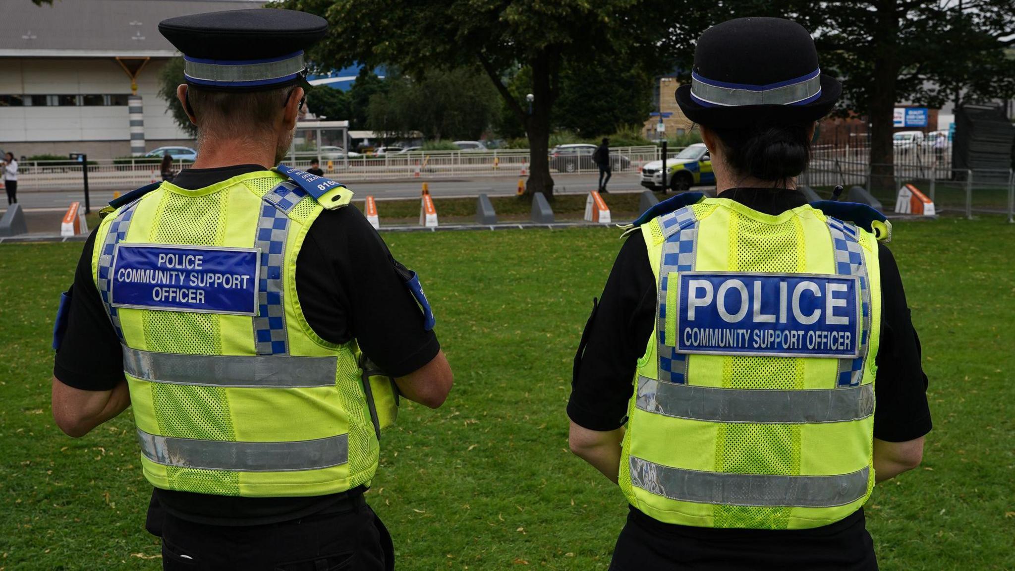 The back of two police community support officers who are stood on grass. Both are wearing a black hat, black t-shirt and a high visibility vest with his job title on