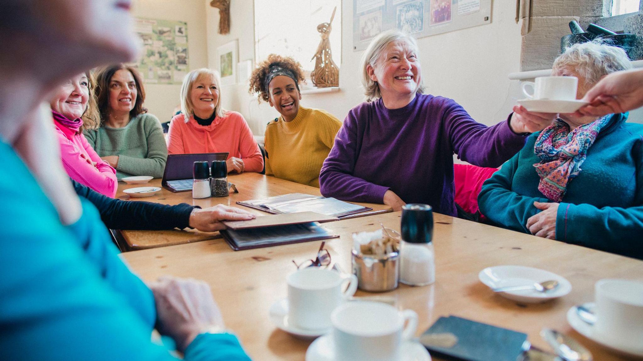 A group of women sit at a table smiling. There are white tea cups on the brown table and everyone is looking to the right of the picture.