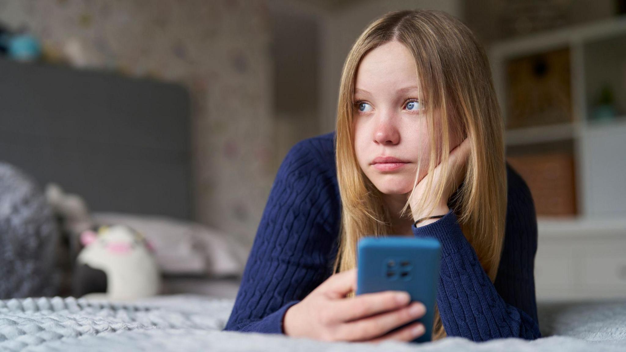 Unhappy Teenage Girl With Mobile Phone Lying On Bed At Home Anxious. She has blonde hair and is wearing a blue jumper and holding a blue phone.