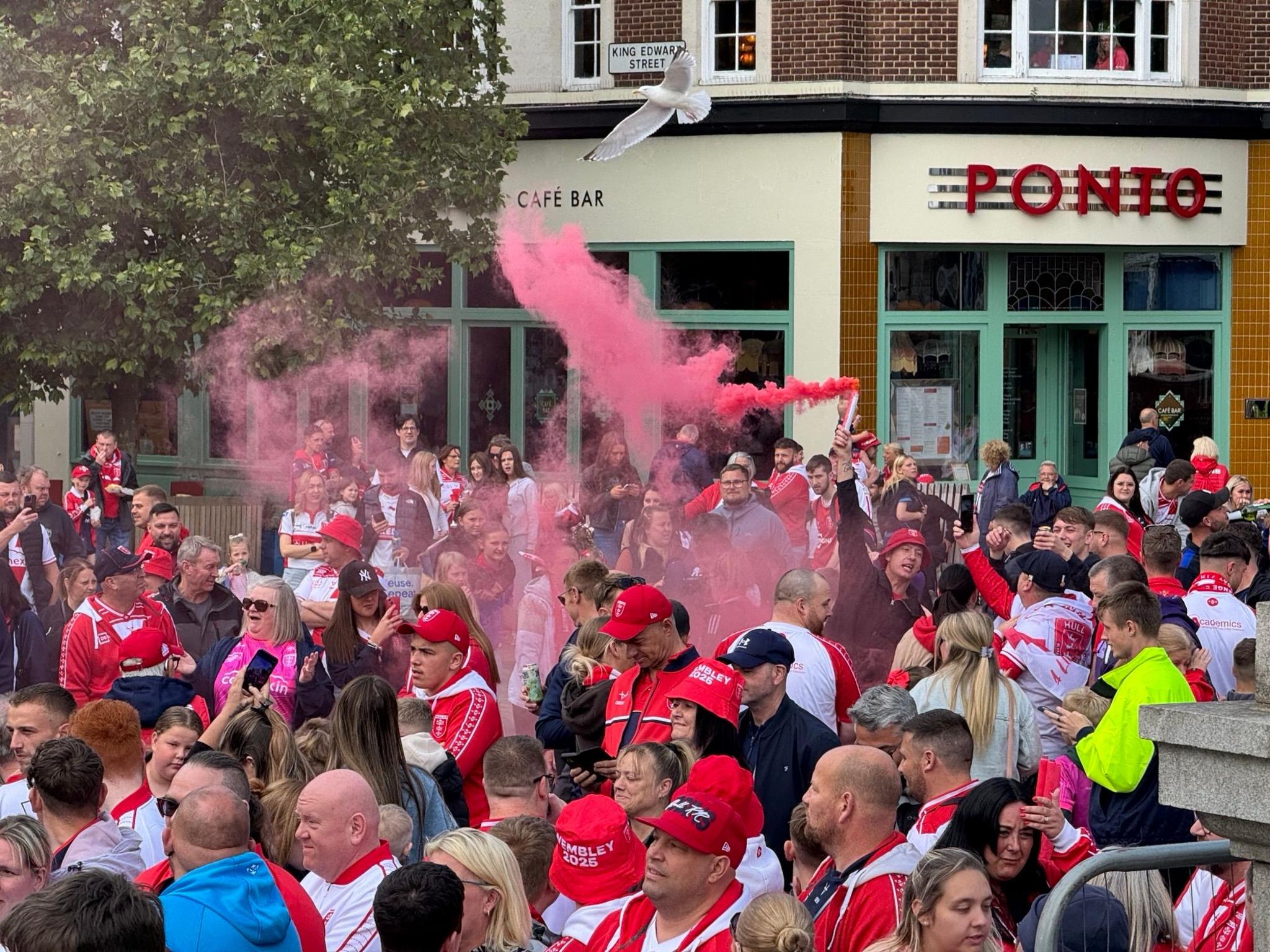 A man holds a flare above his head as it emits red smoke. He is surrounded by a large crowd of people who are wearing red and white clothing.