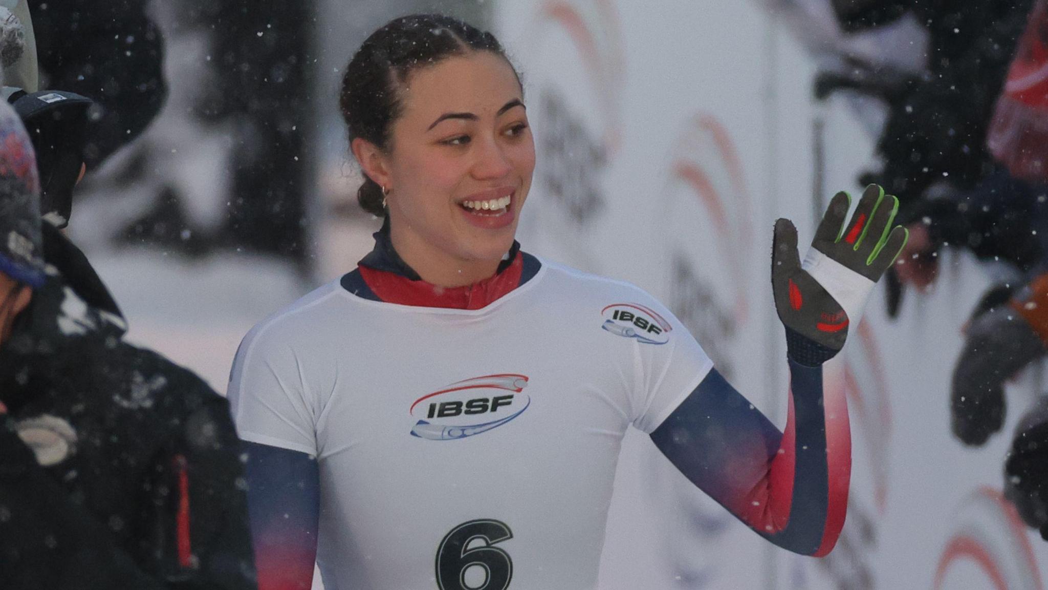 Tabby Stoecker holds her left arm in the air and waves and smiles as she walks on the track following a run in the women's event at the 2026 Winter Olympics