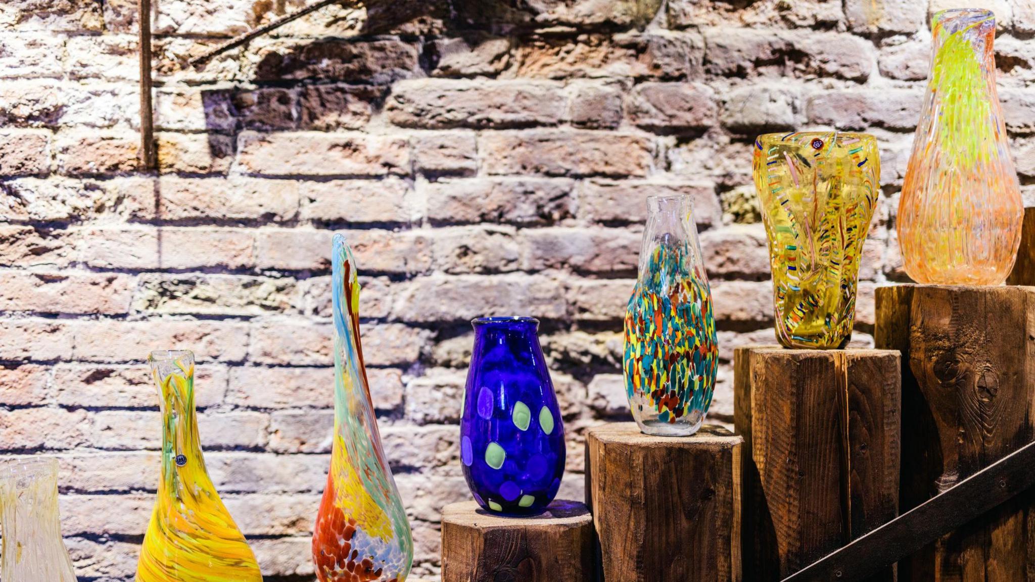 Six colourful Italian Murano glass jugs, lined up on pieces of wood. There is a brick wall in the background.