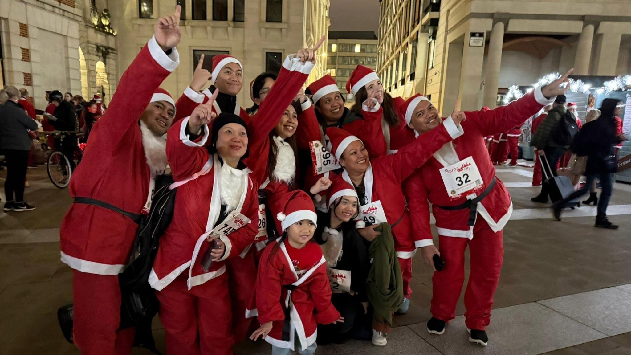 A group of 10 people and one child all posing with their hands in the air and wearing Santa costumes. They're wearing hats and some have white beards around their necks. 