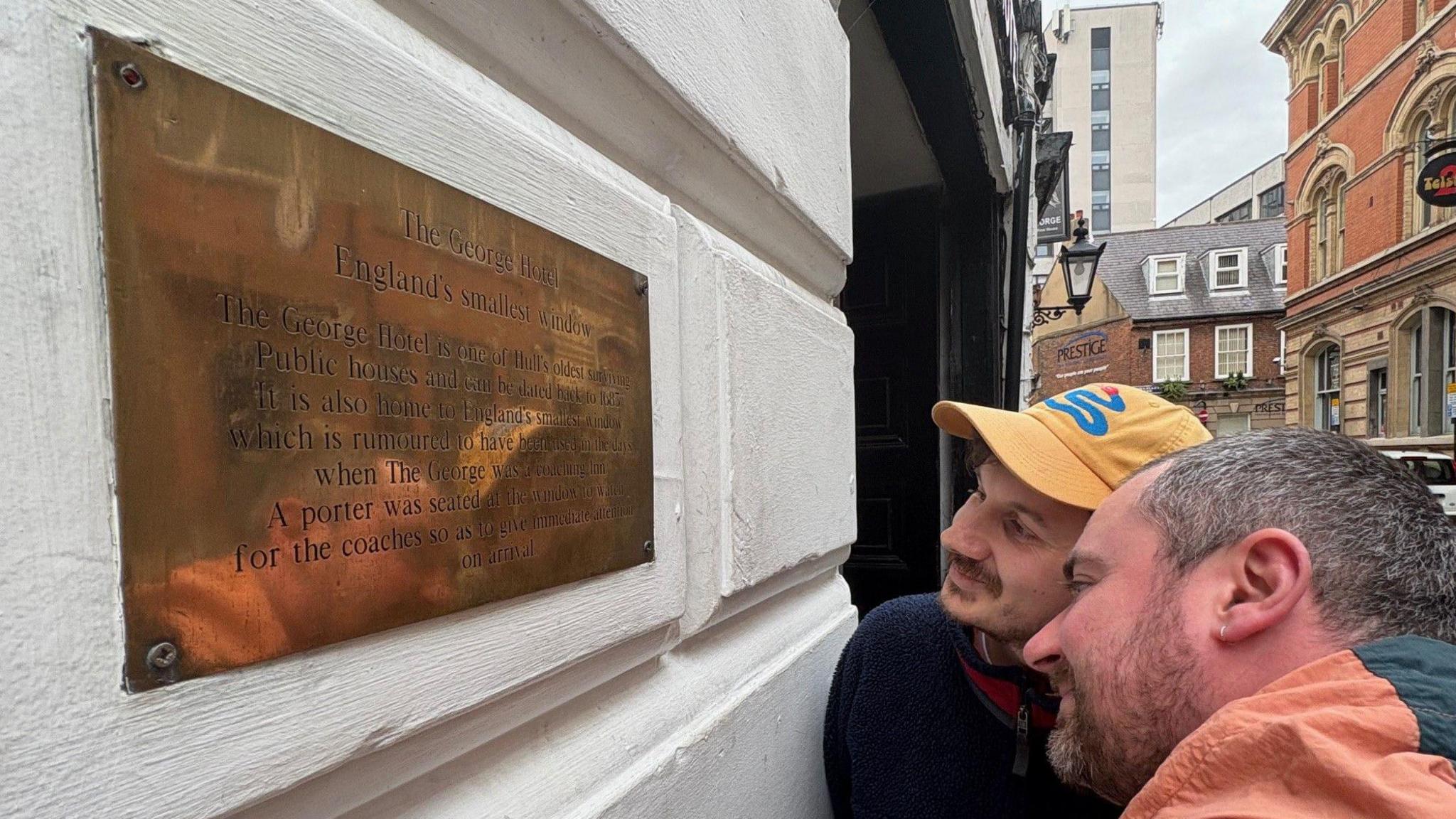 Two men look at a slit window in the site of pub. On the whitewashed wall is a brass plaque claiming the window is the smallest in England.