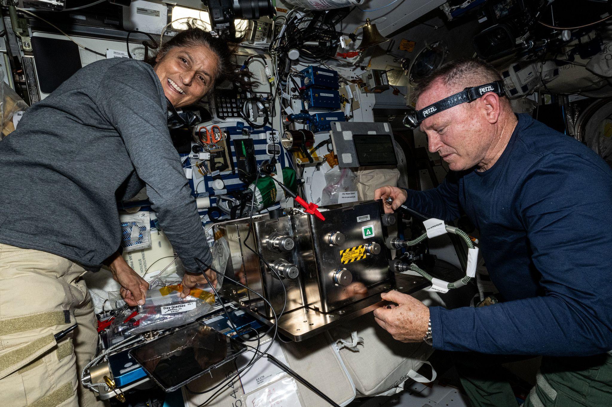 Butch and Suni on ISS - both of them are wearing casual clothing and working on what appears to be complicated equipment with lots of wires and consoles visible. Suni appears to be leaning on one piece of equipment and is smiling, looking over her shoulder at something. Butch has a head torch on and is concentrating on a task.