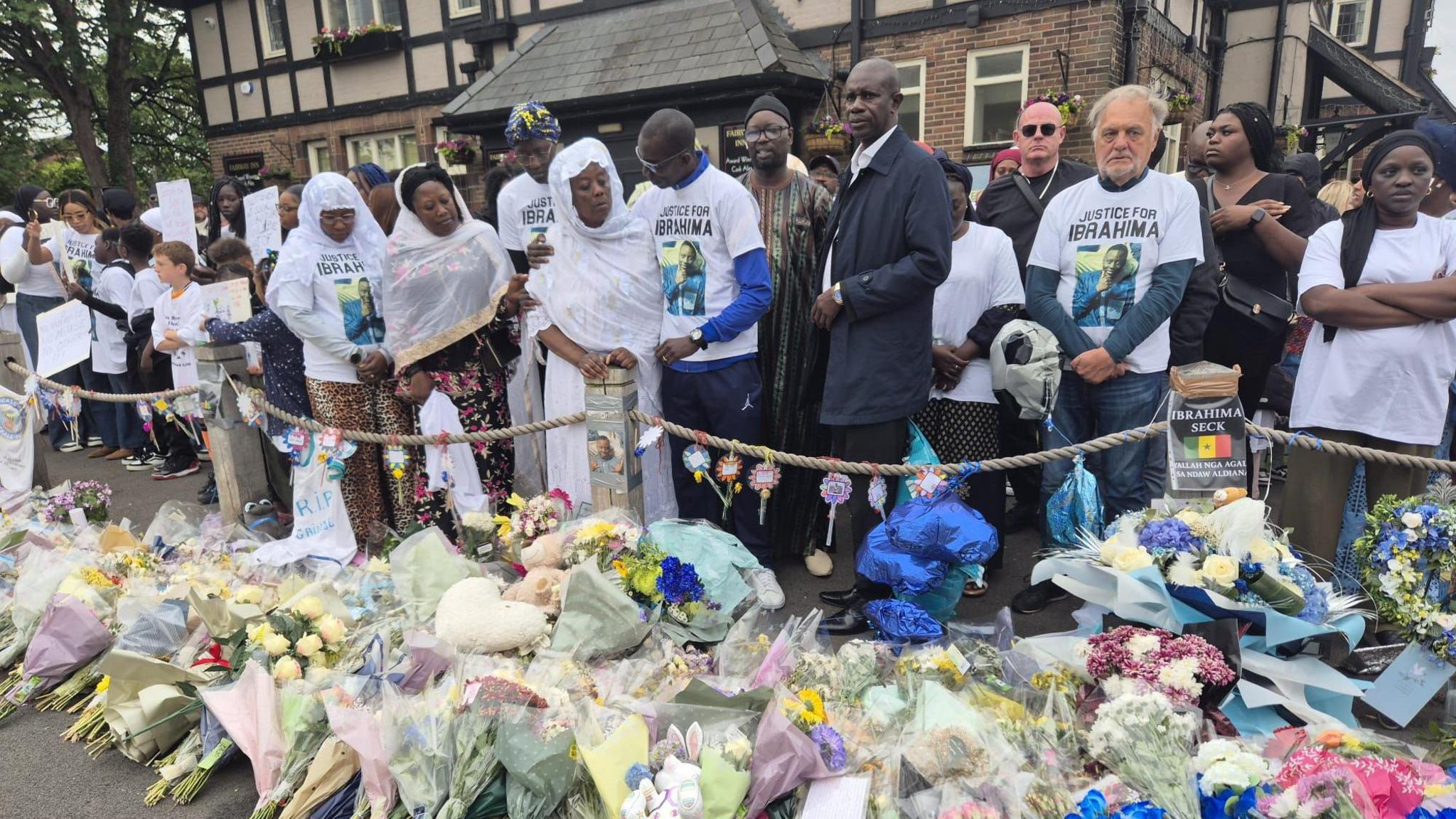Ibrahima Seck's father clasps his arm around his wife while others stand beside them wearing white shirts with Ibrahima's picture. Floral tributes are laid out on the ground in front of them.