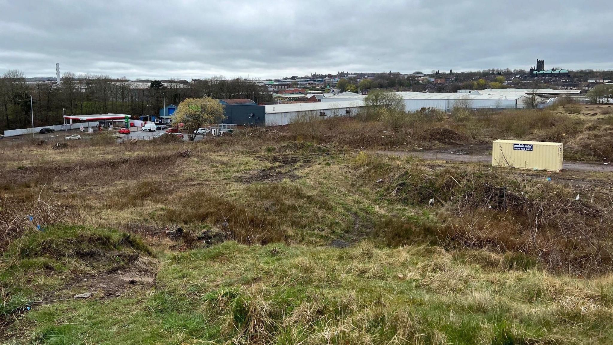 An area of land behind a set of industrial units with a petrol station and Tunstall town centre in the distance.