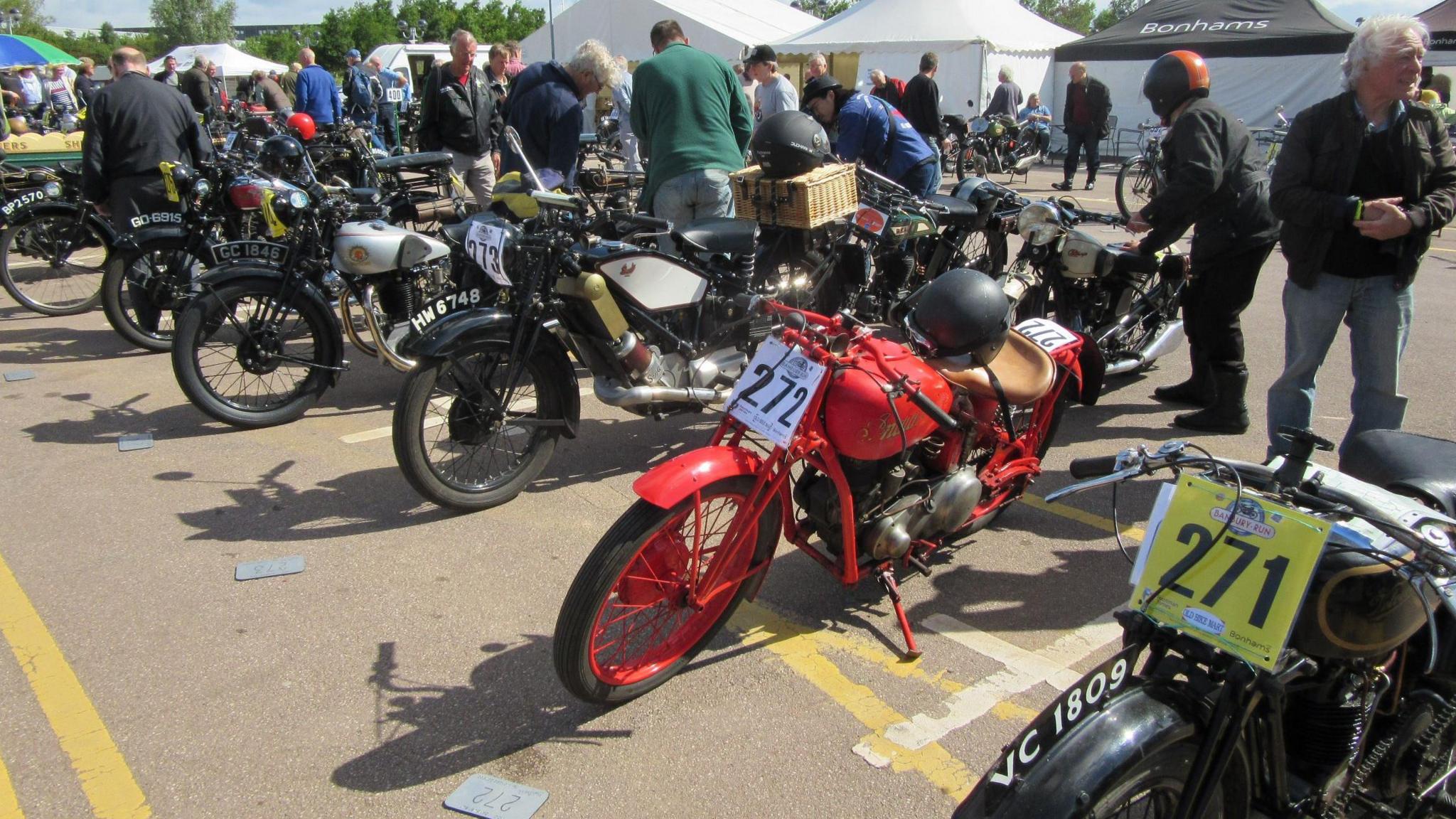 A number of vintage motorcycles, most of them black but one bright red, standing in a line in what looks like a car park. People are milling around and there is a row of gazebos in the background.
