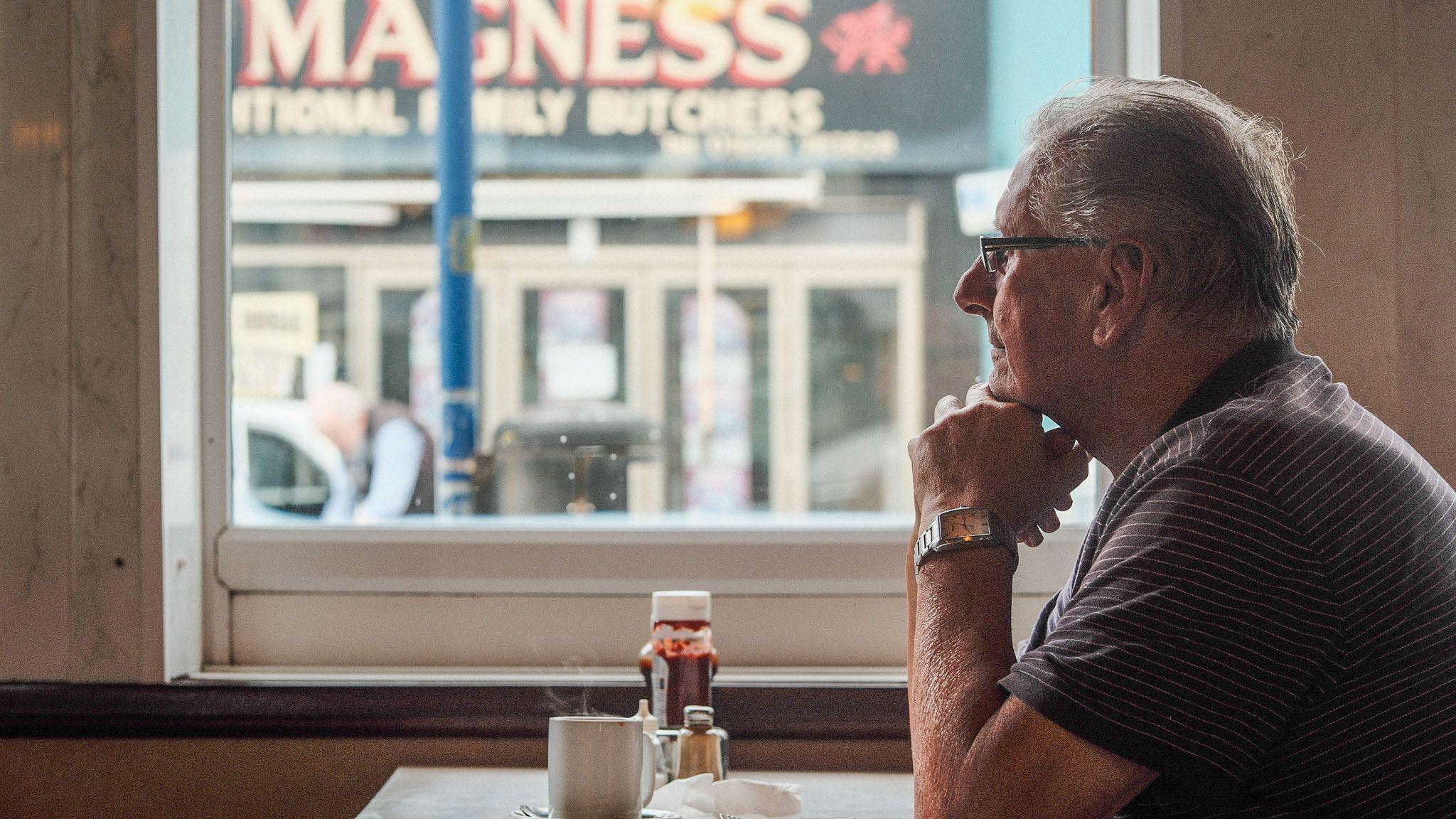 Man sitting in a cafe stall with coffee and another shop with a welsh dragon visible through the cafe window