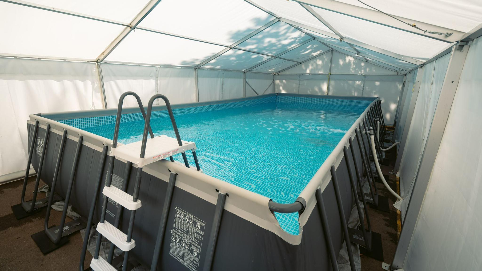 A pool with a grey outer wall and a blue tiled image on the internal wall inside a marquee 