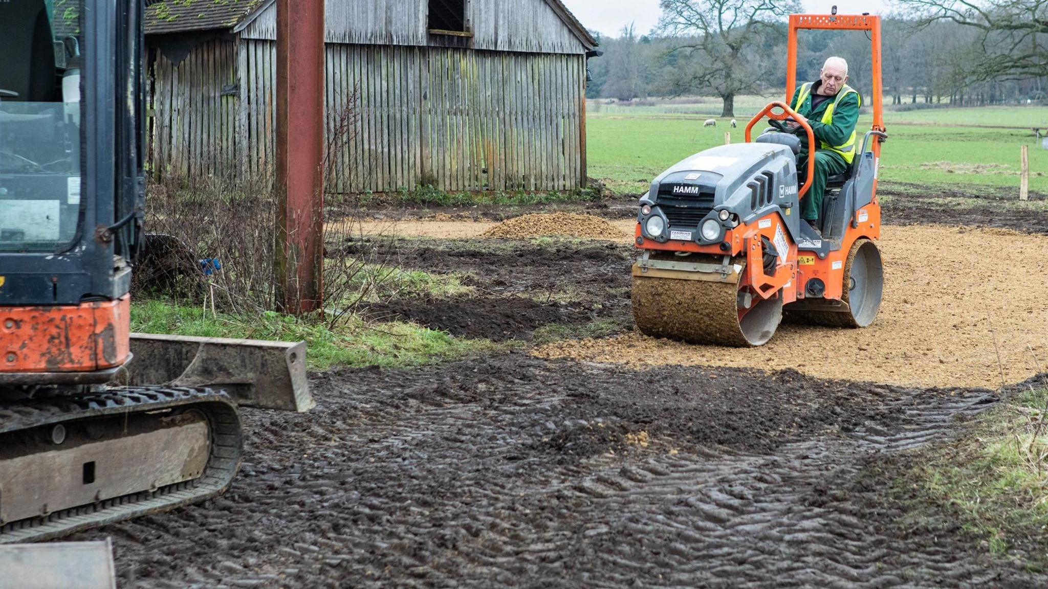 A contractor is driving a road roller at a rural farm site.