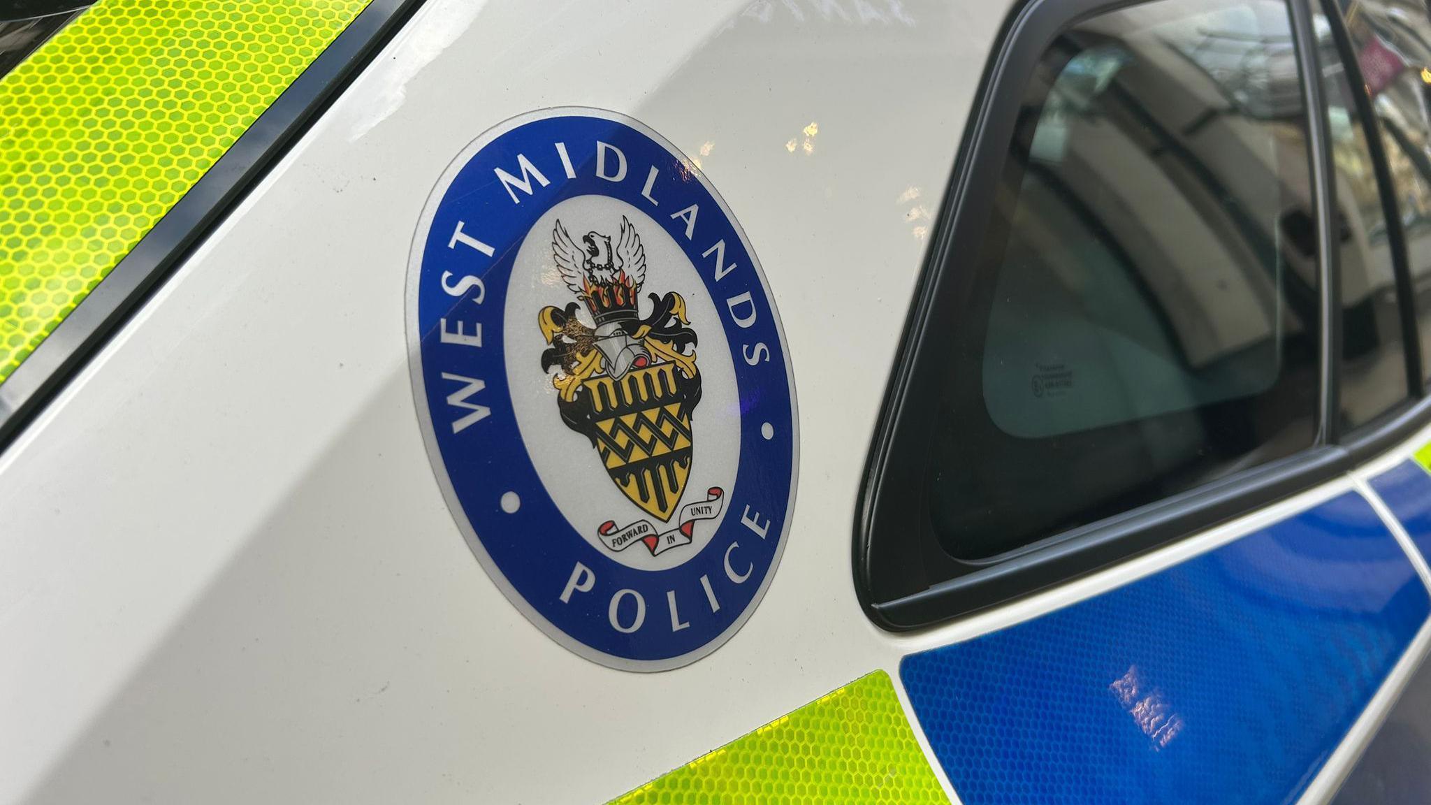 A West Midlands Police emblem stick on the rear part of a white police car in a city centre.