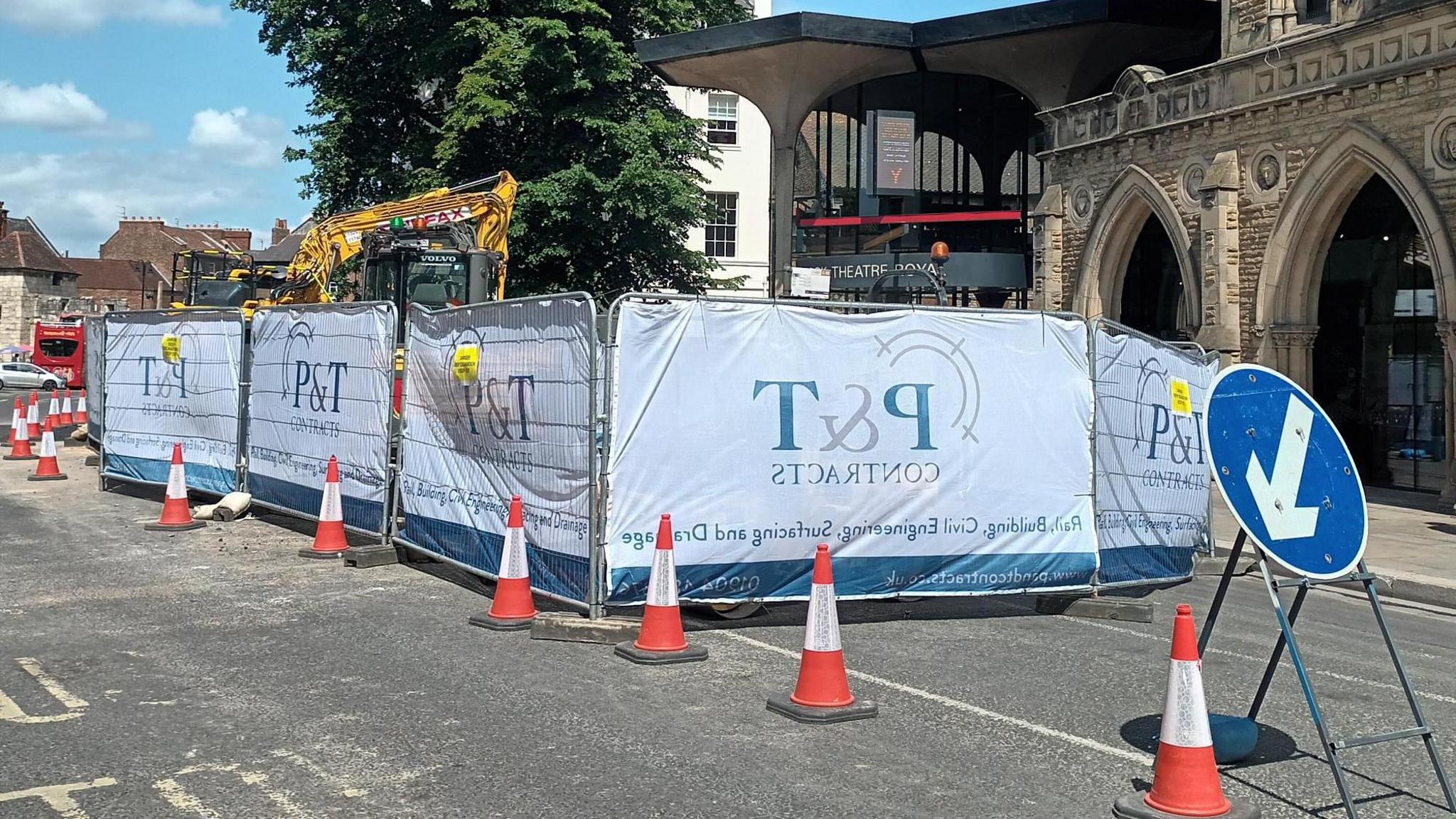 A view of a road, cordoned off with fluorescent orange cones and metal fencing. In the foreground, a blue circular sign can be seen with an arrow directing traffic around the area. In the background a digger can be seen, and a stone building in the background with archways.