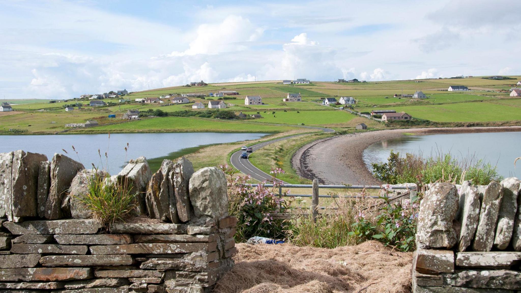 A view over a drystone dyke towards Echna Loch and Echna Bay, Burray. A car crosses a World War Two structure in the loch called a Churchill Barrier. On the island are white-walled houses, almost scattered across a landscape of green fields.