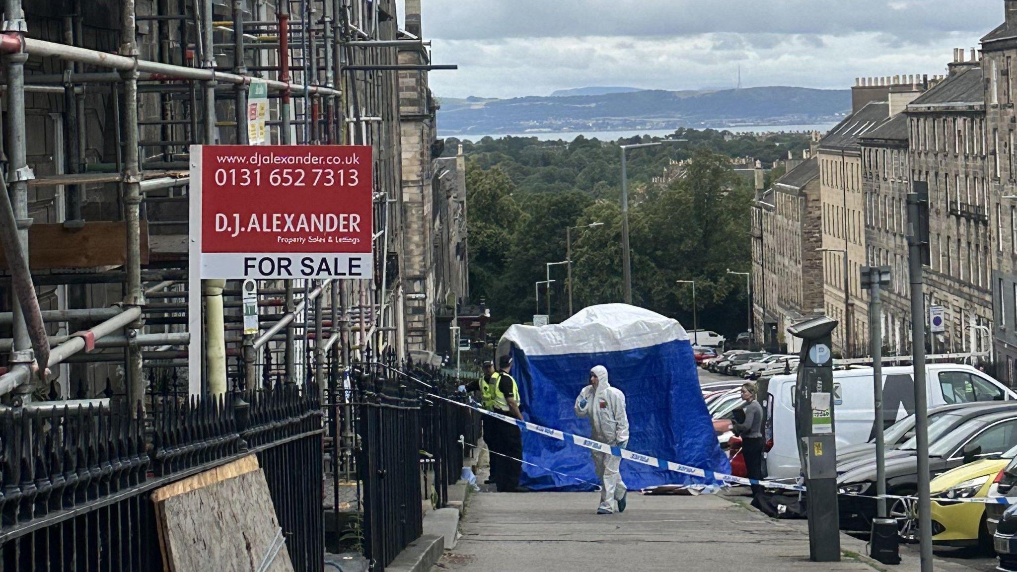 Street showing police tape, a blue police tent and scaffolding against a building