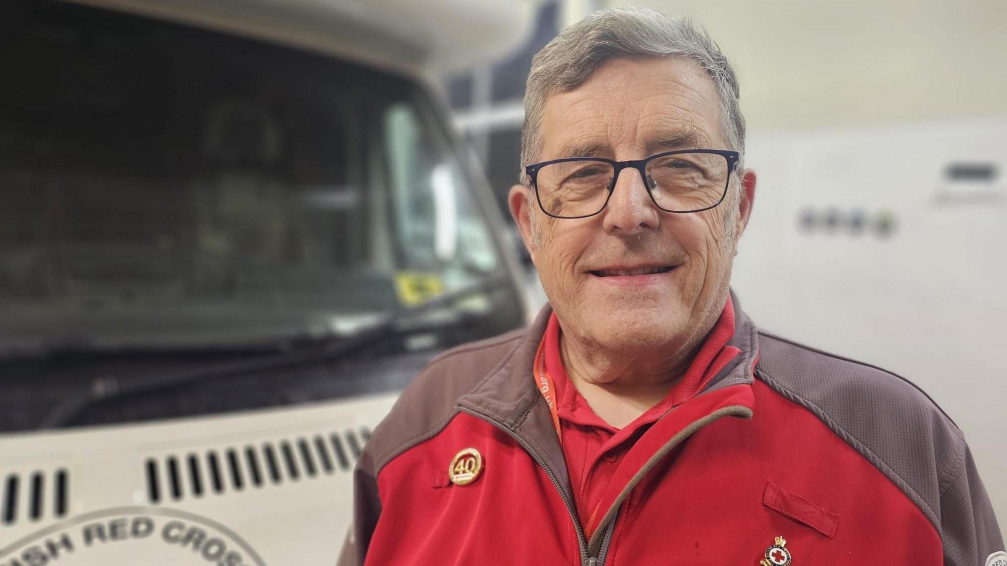 Steve has short grey hair and black rimmed glasses. He is wearing a red jacket with grey shoulders and is smiling at the camera. Behind him is a white camper van.
