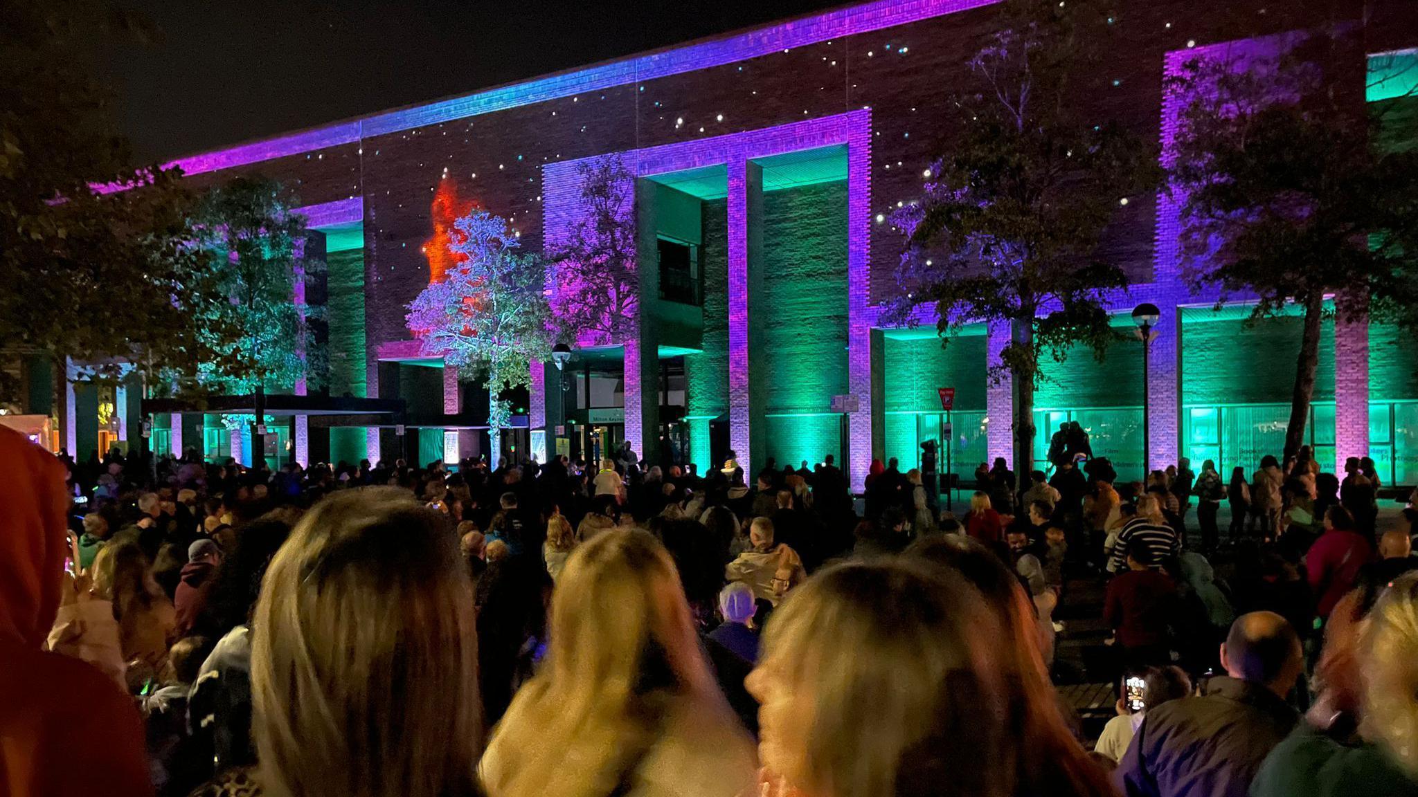 Library Projection City of Codes & Lights 2023, showing crowds of people standing outside a building, with trees, the lights are purple and green. 