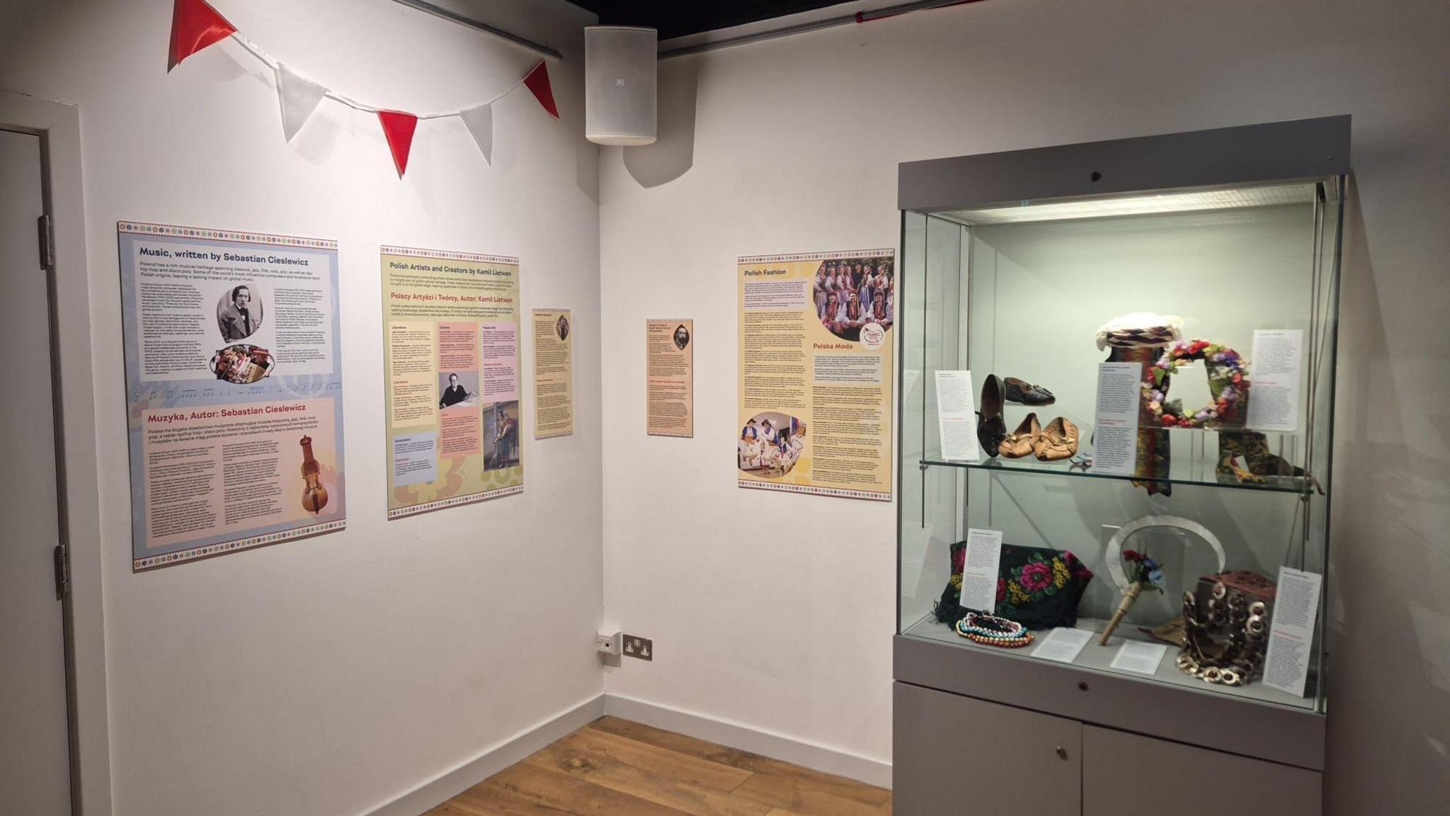 A glass cabinet with artefacts, including shoes and jewellery, highlighting Polish culture. It is standing in a museum with posters and information boards on a crisp white wall.