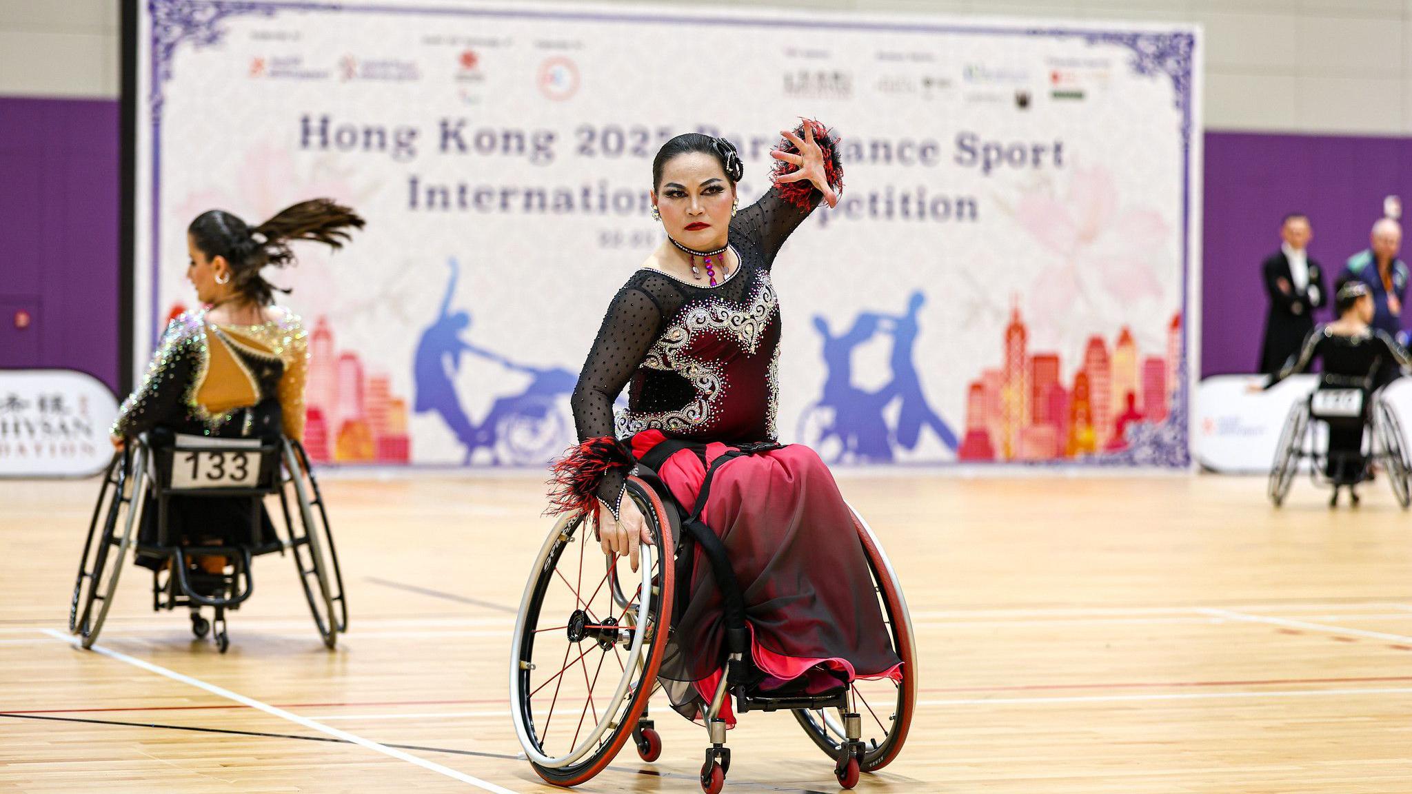 A woman in a dramatic black and red dress with tassles on the cuffs dances in a wheelchair on a wooden floor. She looks serious and has dramatic, dark eye makeup. Behind her a banner reads 'Hong Kong 2025 Para Dance Sport International Competition'. 