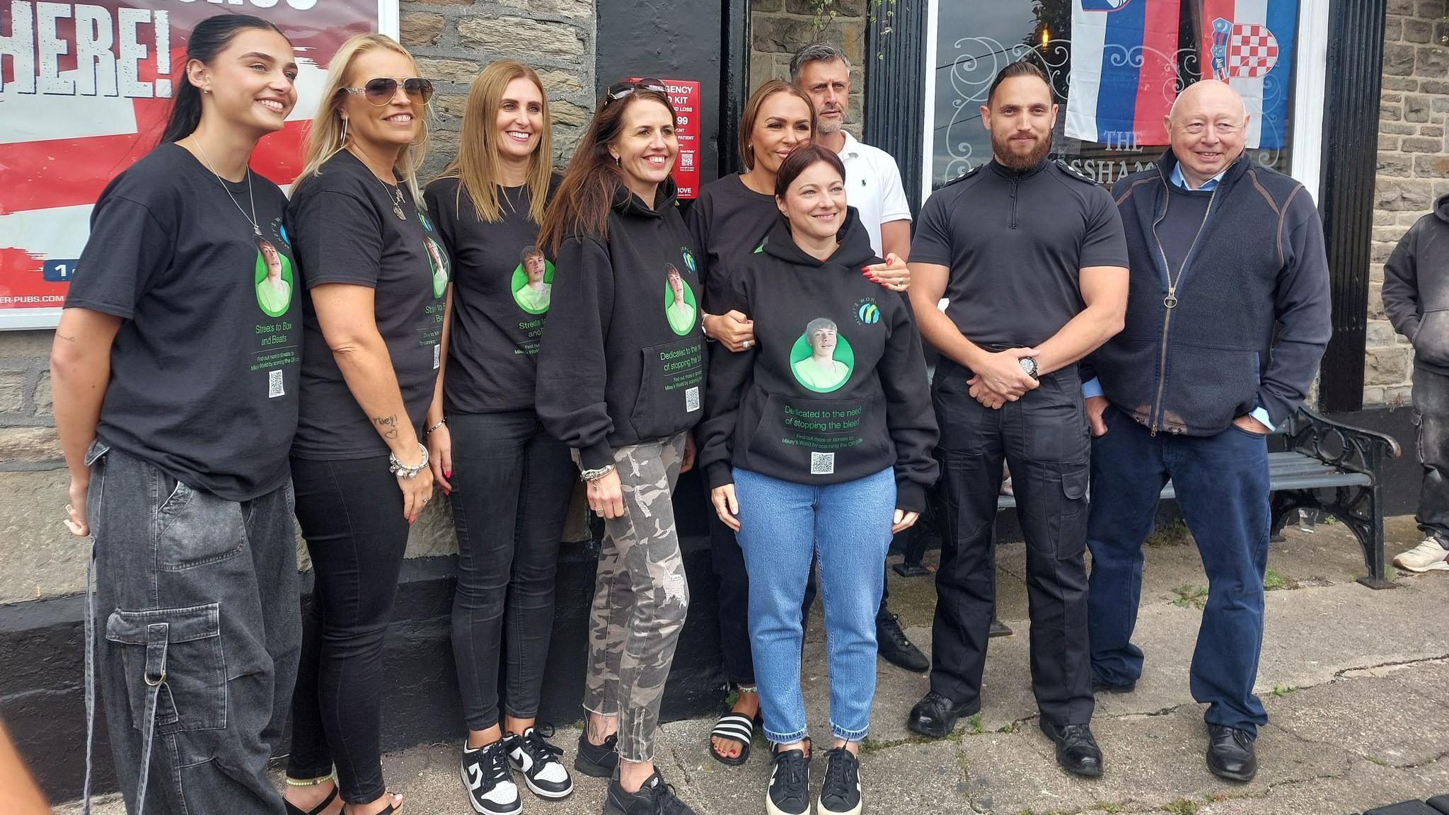 A group of people wearing clothing with Mikey's image on are posing for a photography near a bleed kit. Chief Inspector Mike Vass is also there.