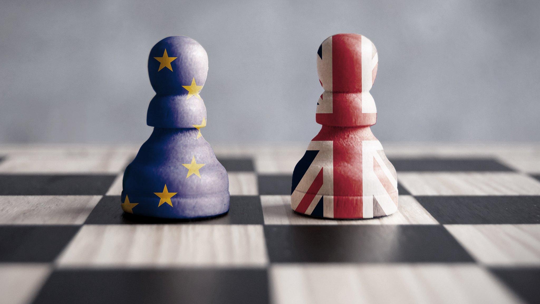 Two chess pieces on a chess board, one enclosed in the EU flag, one enclosed in the Union flag.