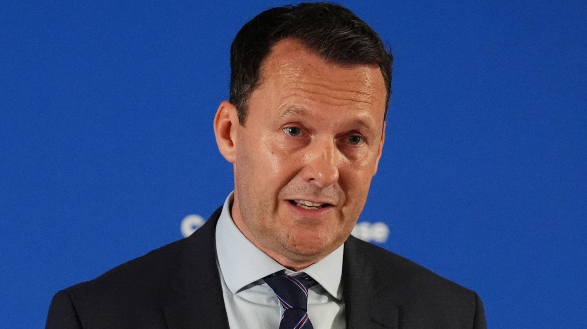 Russell Findlay wears a black blazer and grey shirt and is standing in front of a blue backdrop