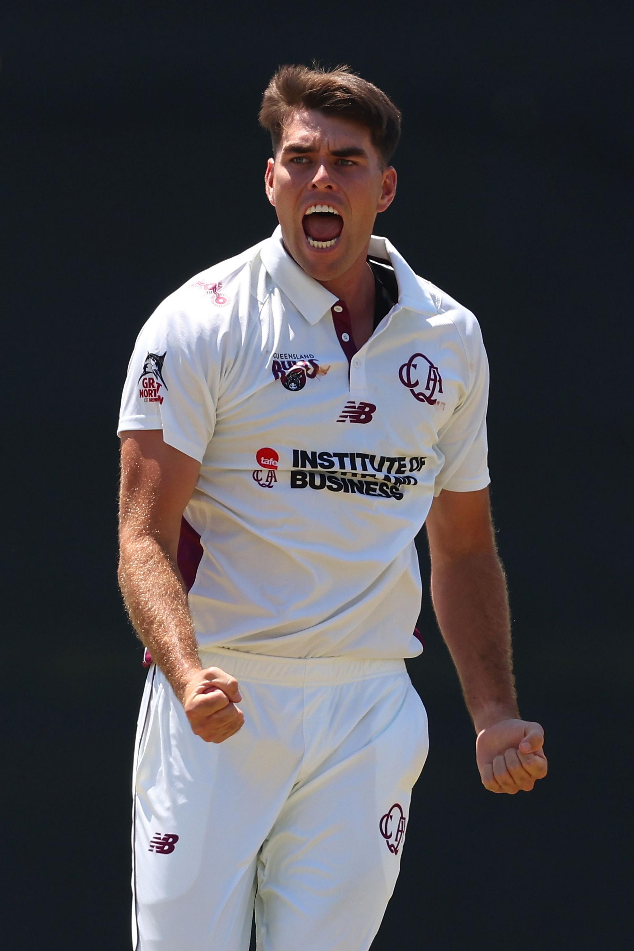 Cricket player in a white uniform with maroon accents, clenching both fists in a celebratory gesture, standing against a dark background.
