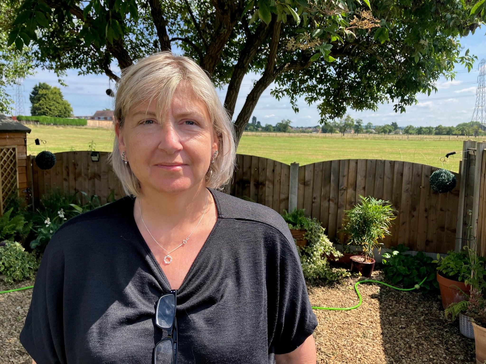 A woman with chin-length blonde-grey hair in a bob stands in a garden on a sunny day. A fence behind her looks out over a large field.