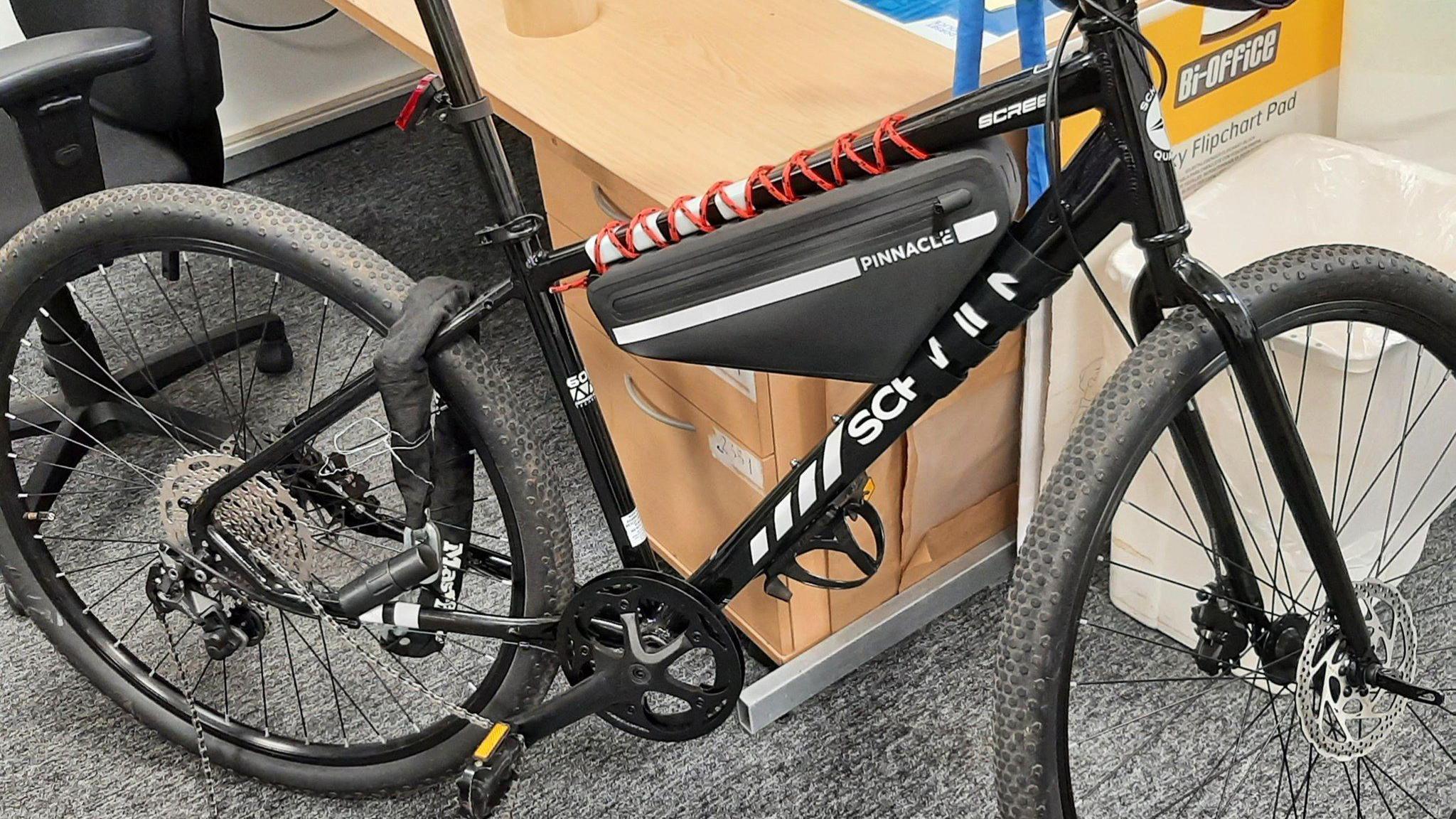 A black bike leaning against a desk in an office with a grey carpet. It still has a lock attached to the back wheel.