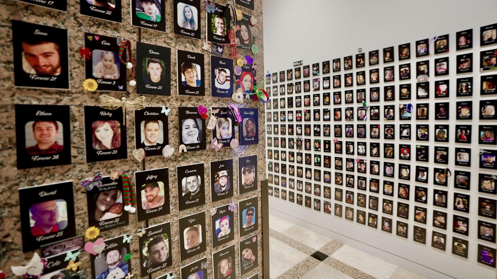 Two walls which are covered with the names and images of people who have died, tightly packed so there are 10 pictures in each column - and the columns continue beyond the edge of this photograph. Below each picture is the age the person died at: "Forever 23", for example. Some of the images are decorated with hearts and flowers.